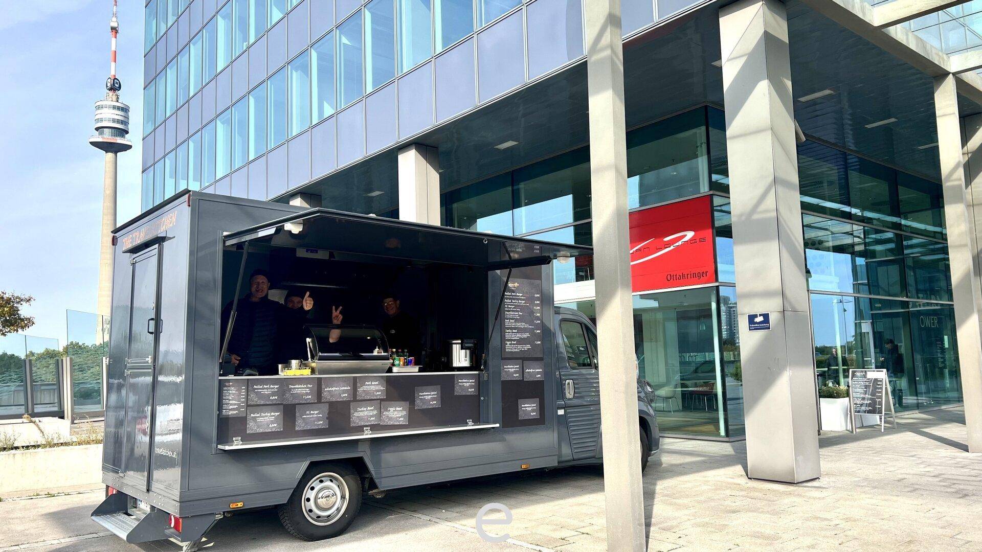 Foodtruck vor einem modernen Bürogebäude mit Glasfassade und einem hohen Turm im Hintergrund.