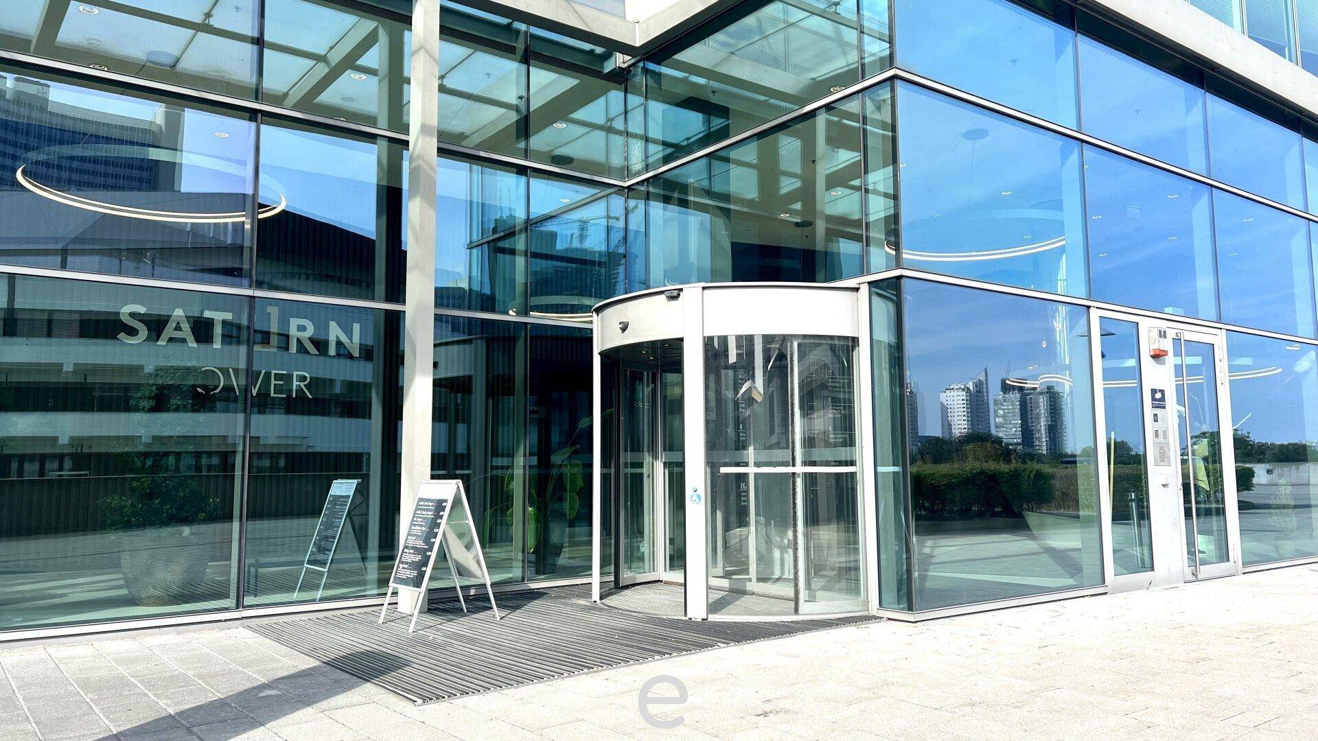 Eingangsbereich eines modernen Bürogebäudes mit Glasfassade und Drehtür unter blauem Himmel.