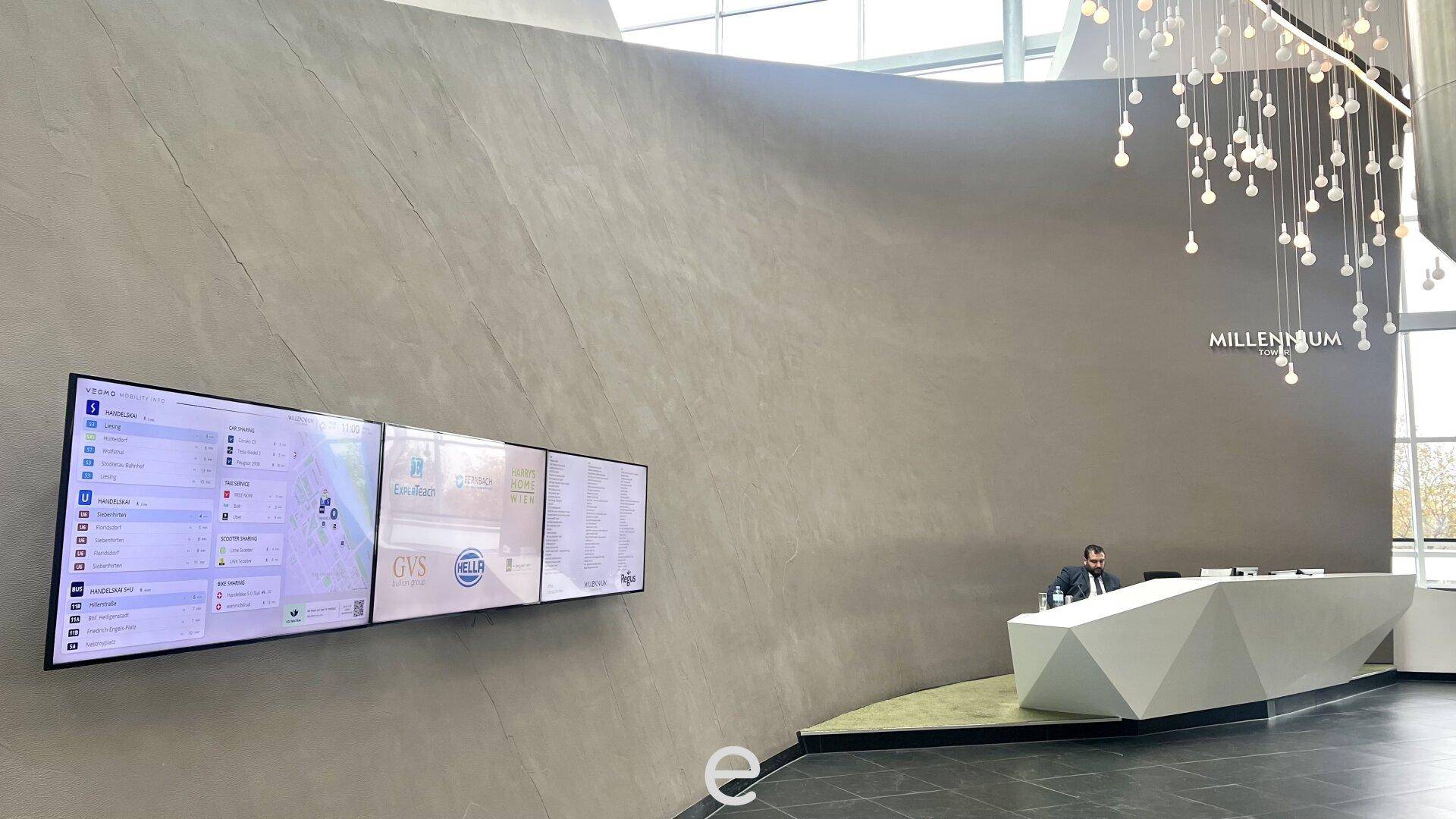 Moderner Empfangsbereich mit geometrischem Schreibtisch, großen Informationsbildschirmen und auffälliger Deckenleuchte.