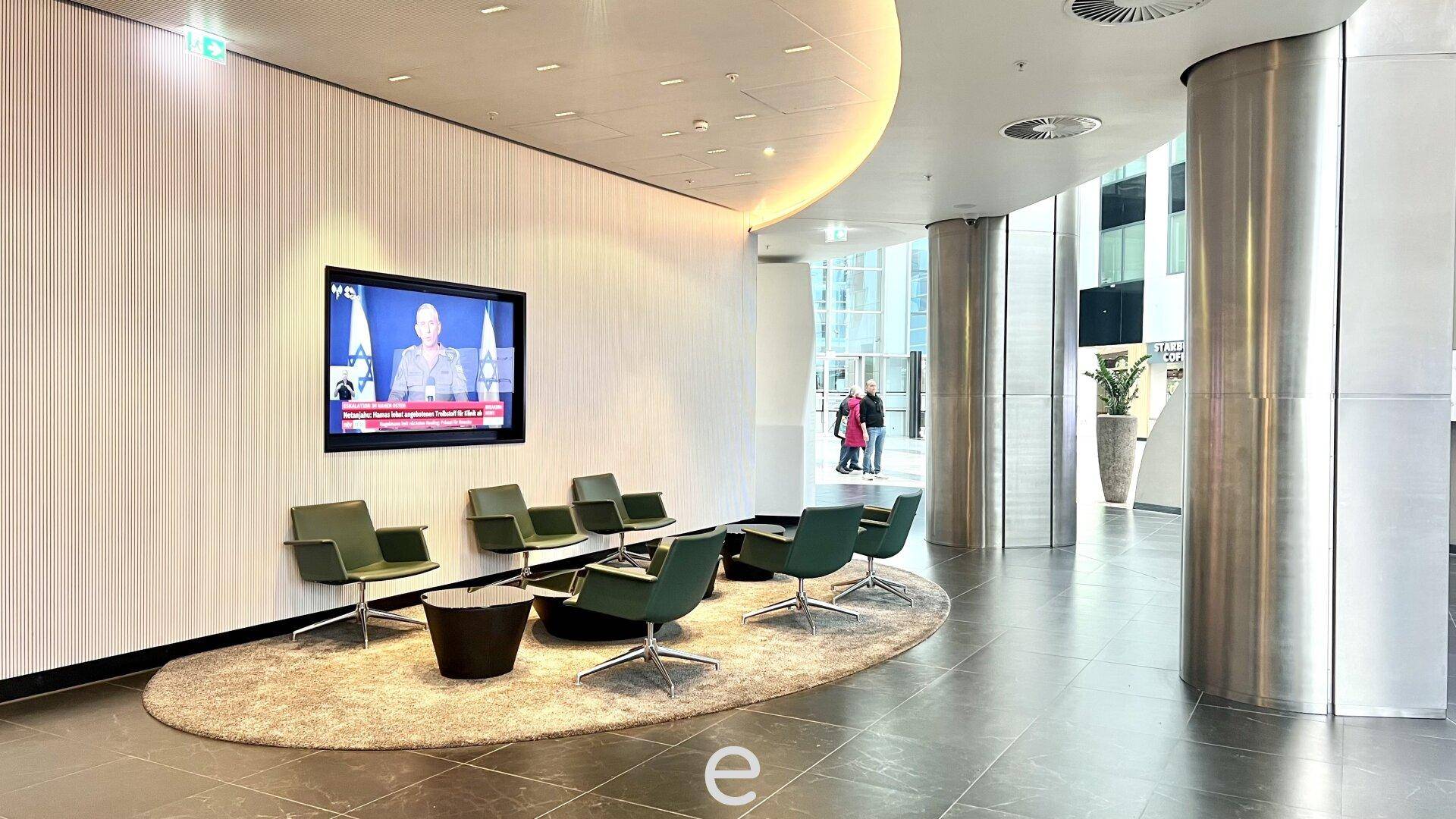 Gemütlicher Wartebereich in der Lobby mit modernen grünen Sesseln, Teppich und großem Bildschirm.