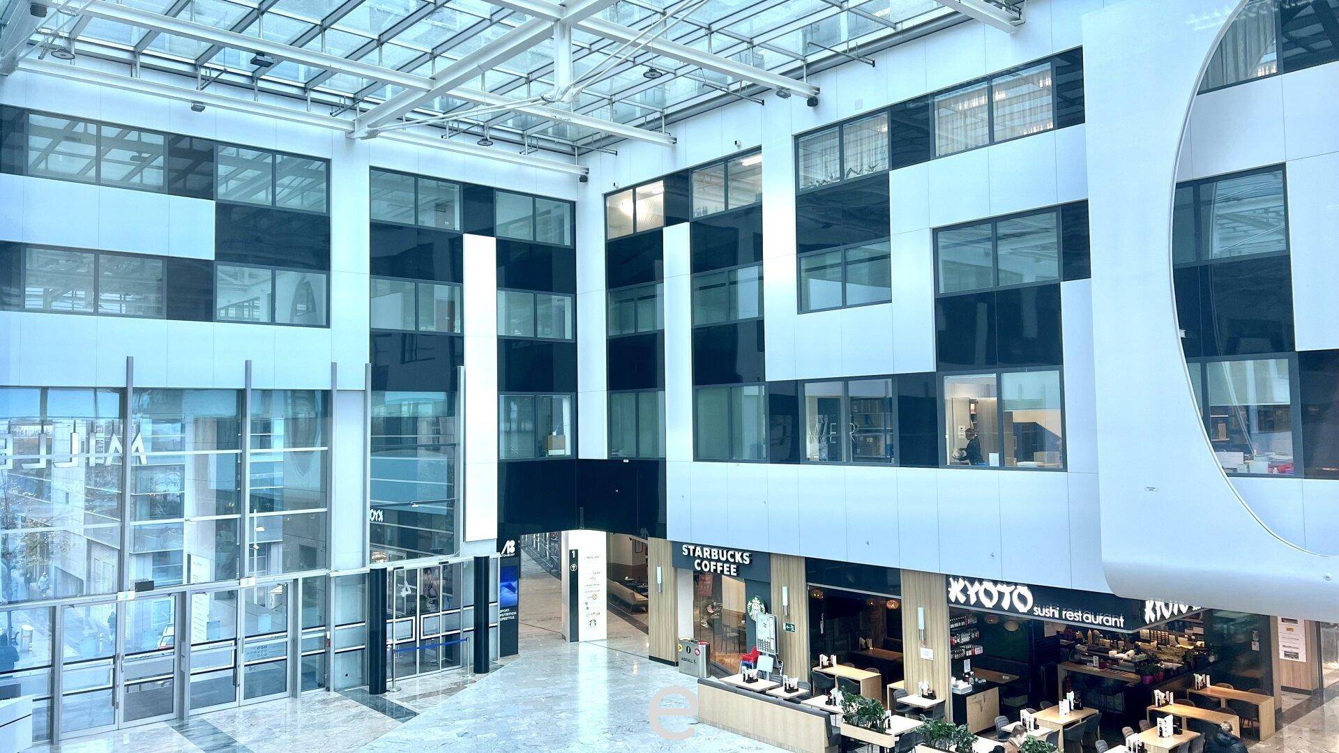 Mehrstöckiges Atrium eines modernen Gebäudes mit Glasfassaden, Geschäften und viel natürlichem Licht.