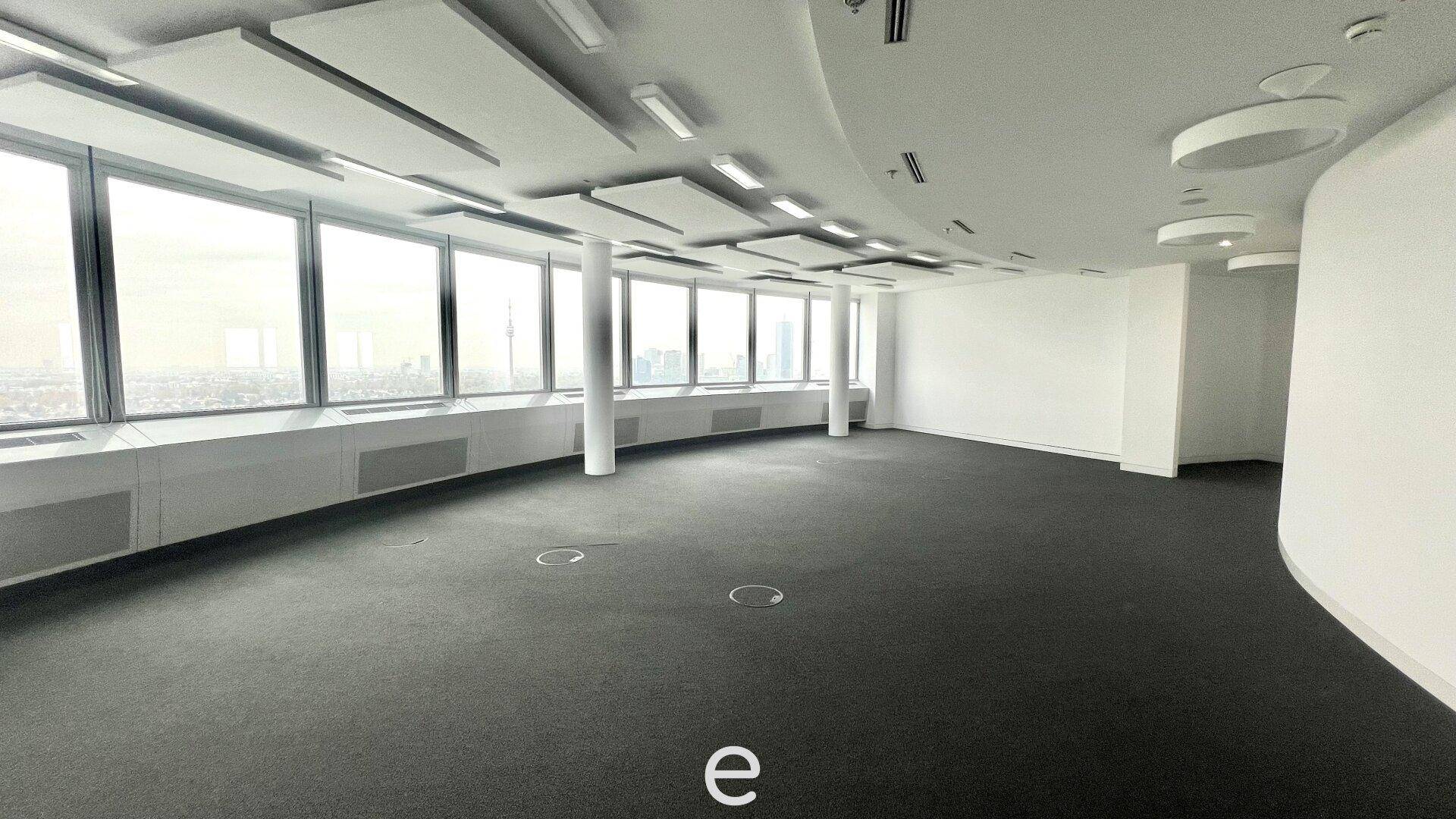 Geräumiger, leerer Büroraum mit Teppichboden, geschwungenen Fenstern und Panoramablick auf die Stadt.