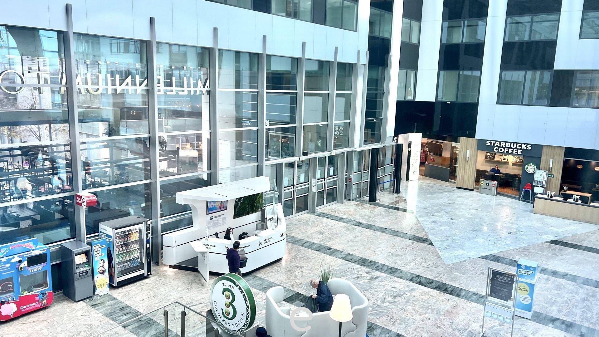 Großes Atrium eines modernen Gebäudes mit mehreren Ebenen, Geschäften wie Starbucks und einem zentralen Informationsschalter.