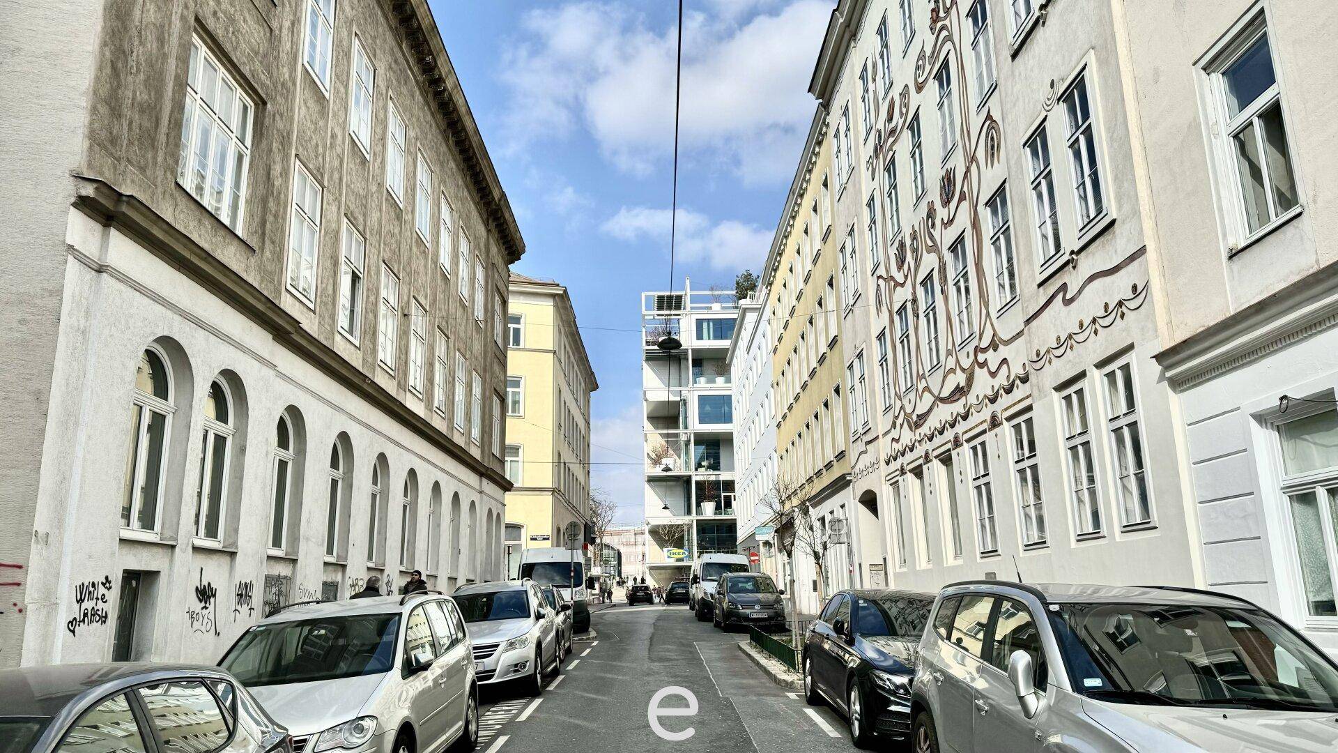 Eine belebte Stadtstraße mit historischen Gebäuden, geparkten Autos und einem modernen Gebäude im Hintergrund.