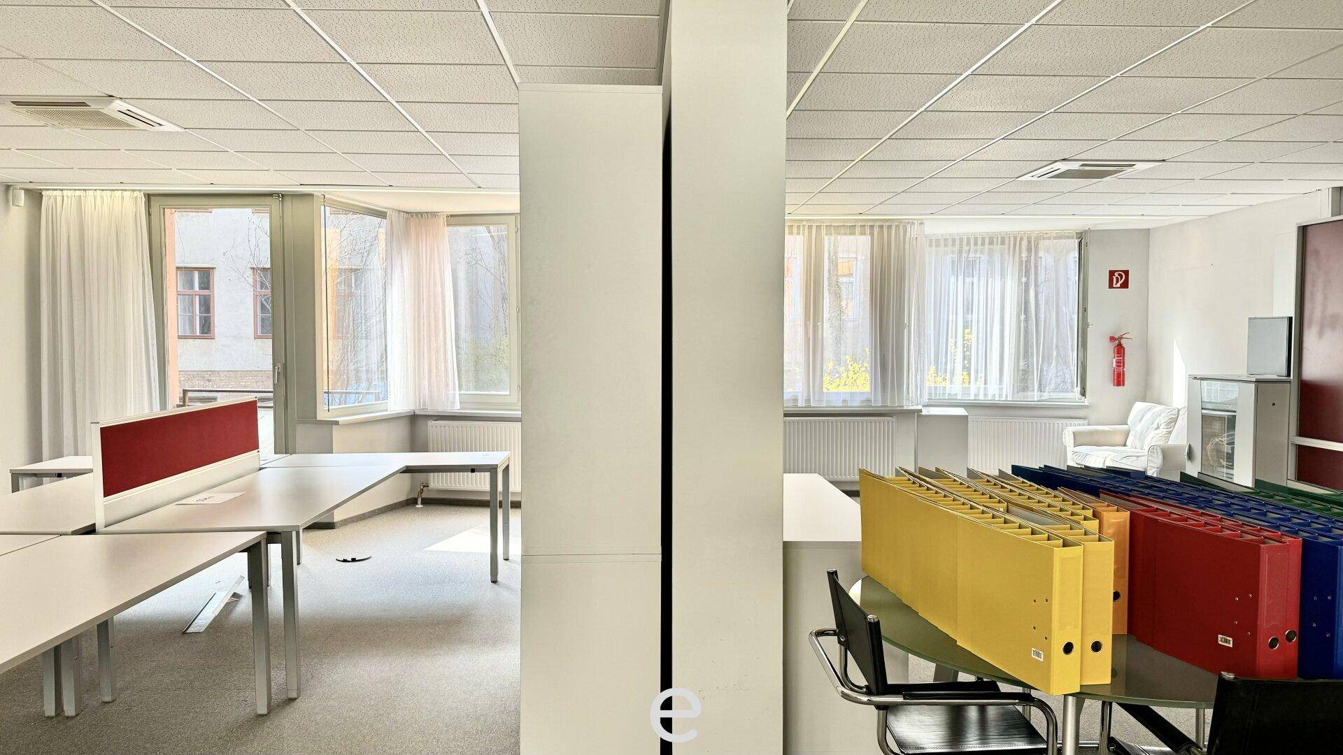 Geräumiges Großraumbüro mit mehreren Arbeitsplätzen, Teppichboden und großen Fenstern für viel Tageslicht.