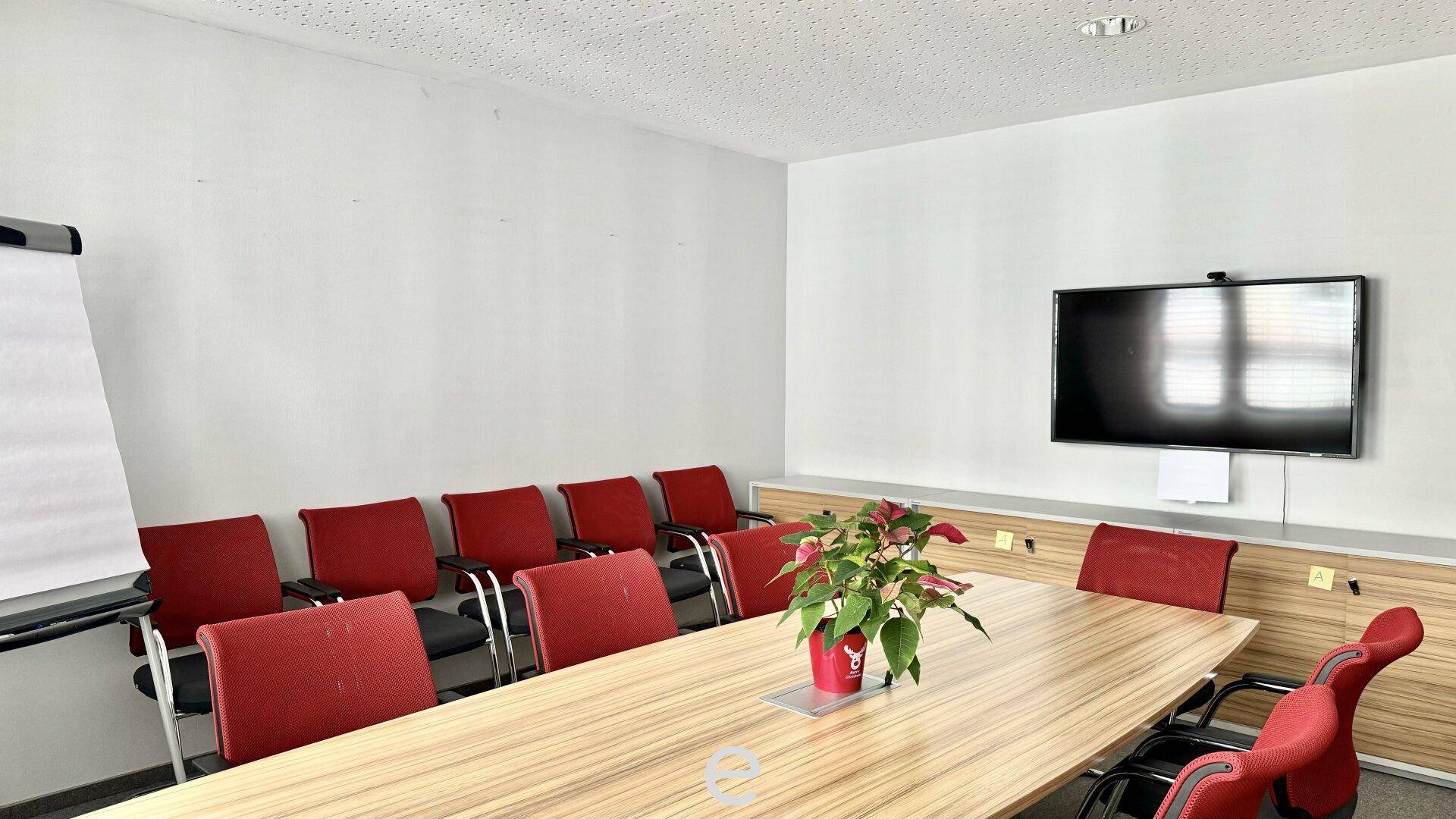 Konferenzraum mit großem Holztisch, roten Stühlen und einem an der Wand montierten Fernseher für Präsentationen.