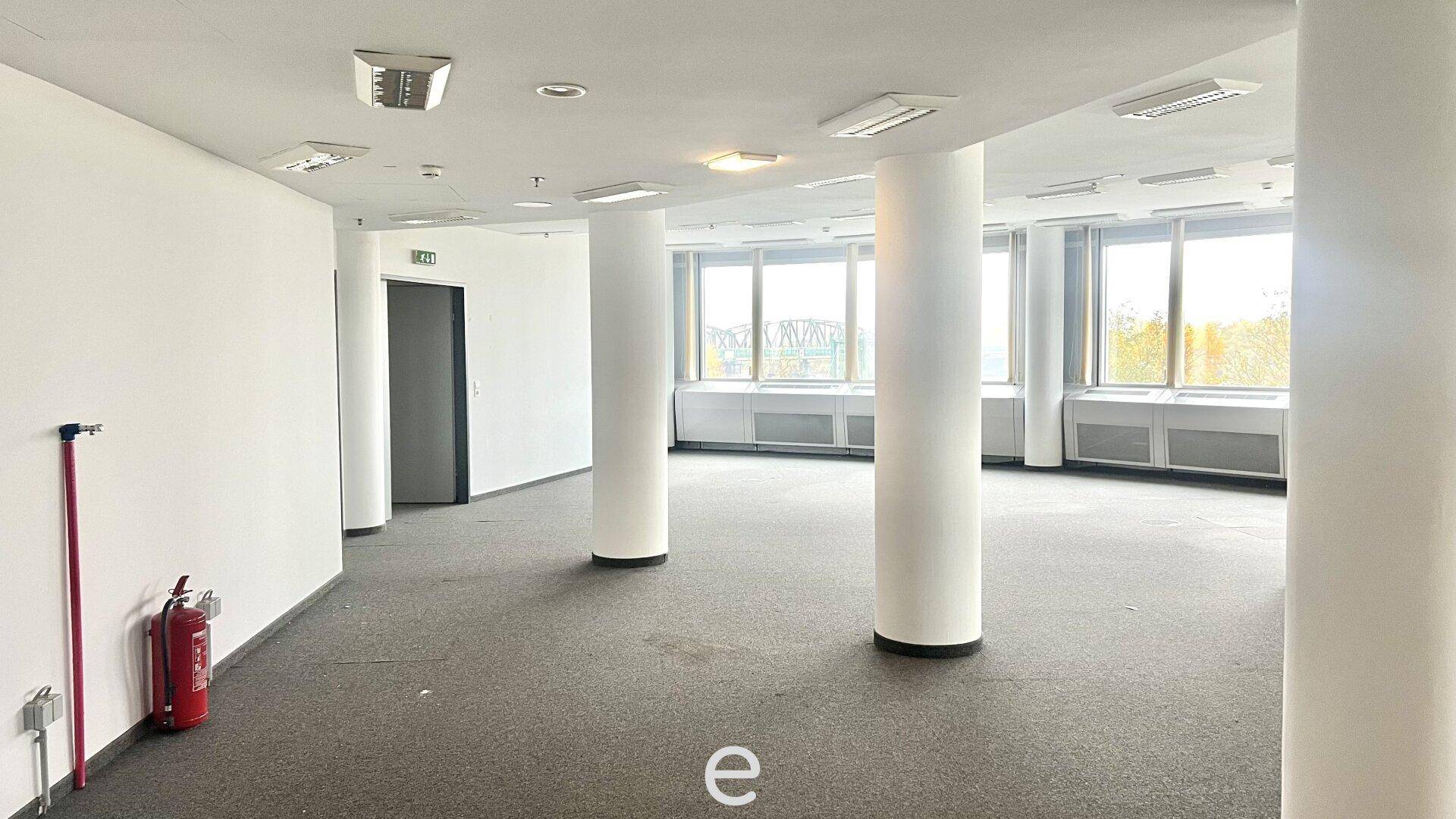 Großes, leeres Büro mit Säulen, Teppichboden und vielen Fenstern, die viel Tageslicht hereinlassen.