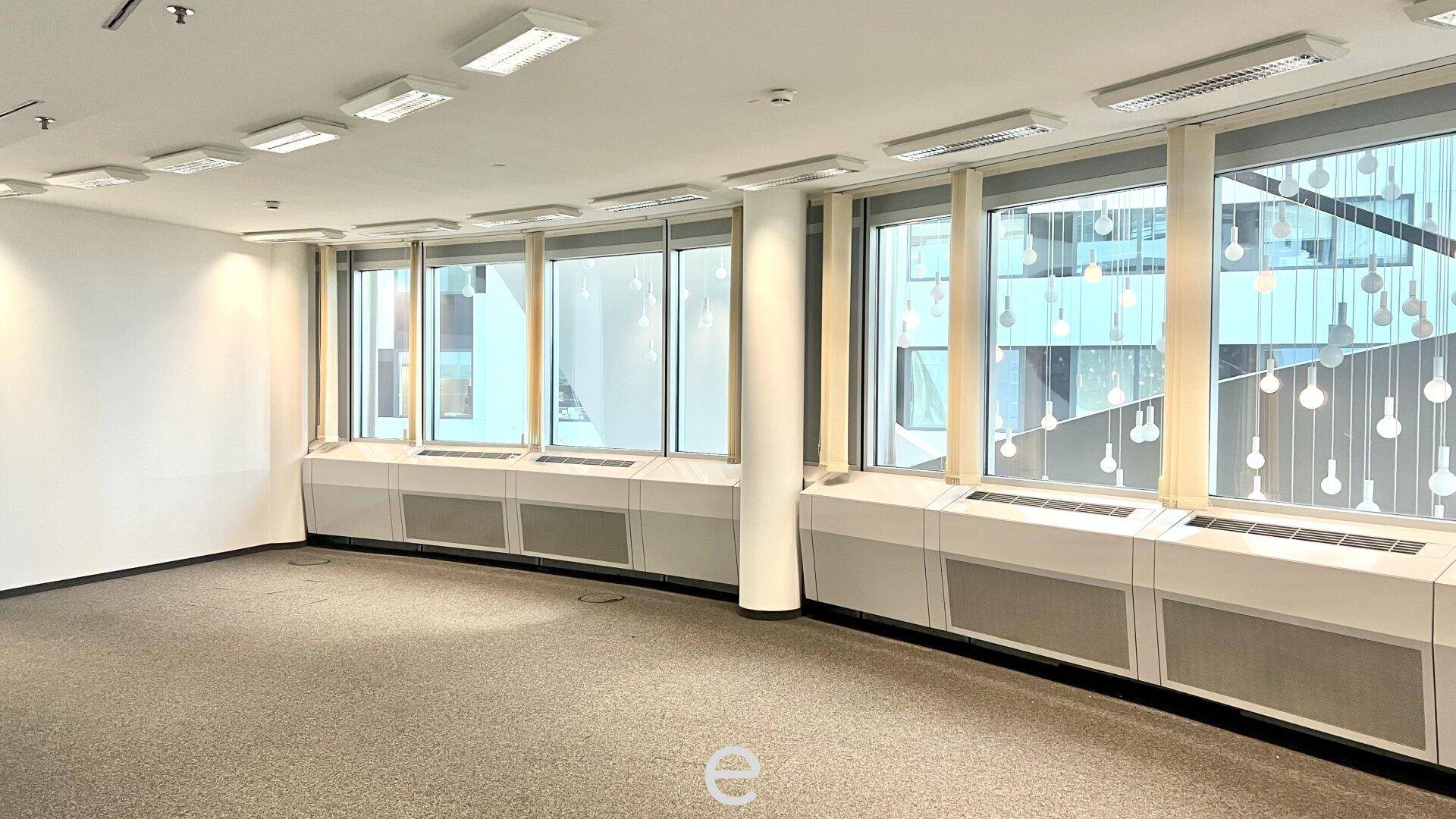 Leeres Büro mit Teppichboden, großen Fenstern und Heizkörpern unter den Fensterbänken.