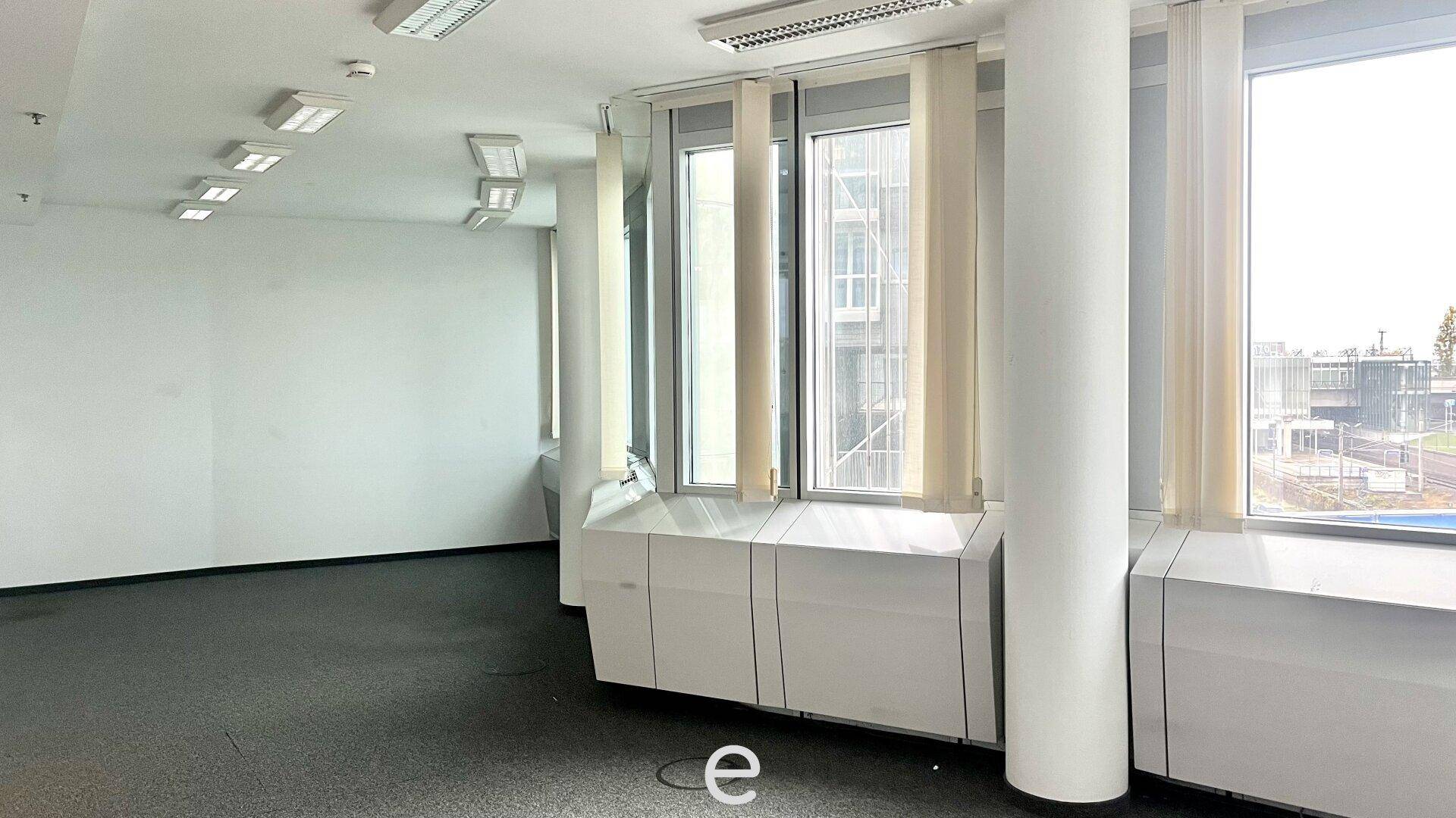 Leeres Büro mit Teppichboden, großen Fenstern und Heizkörpern unter den Fensterbänken.