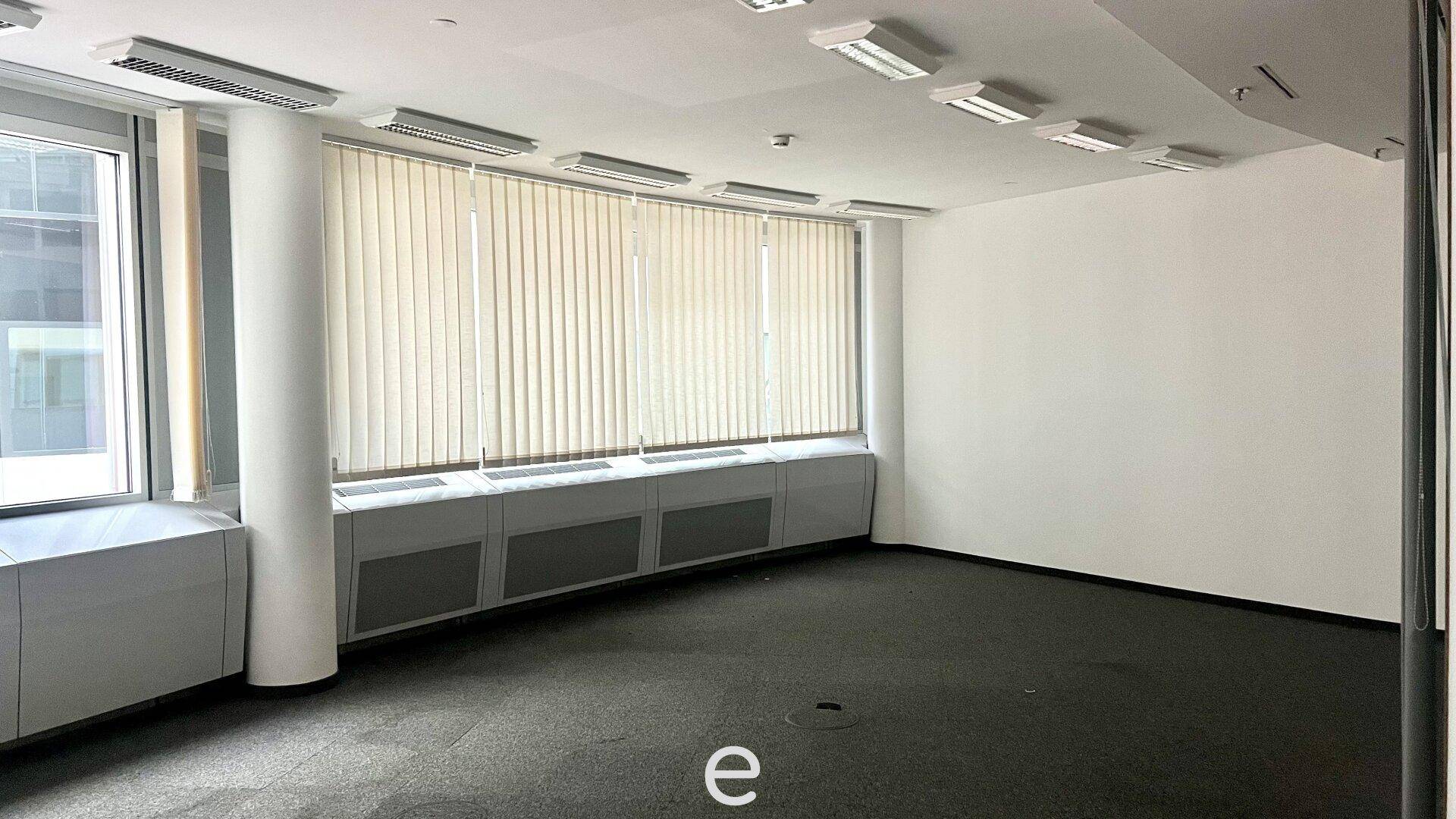 Leeres Büro mit Teppichboden, großen Fenstern und Jalousien für Sonnenschutz.