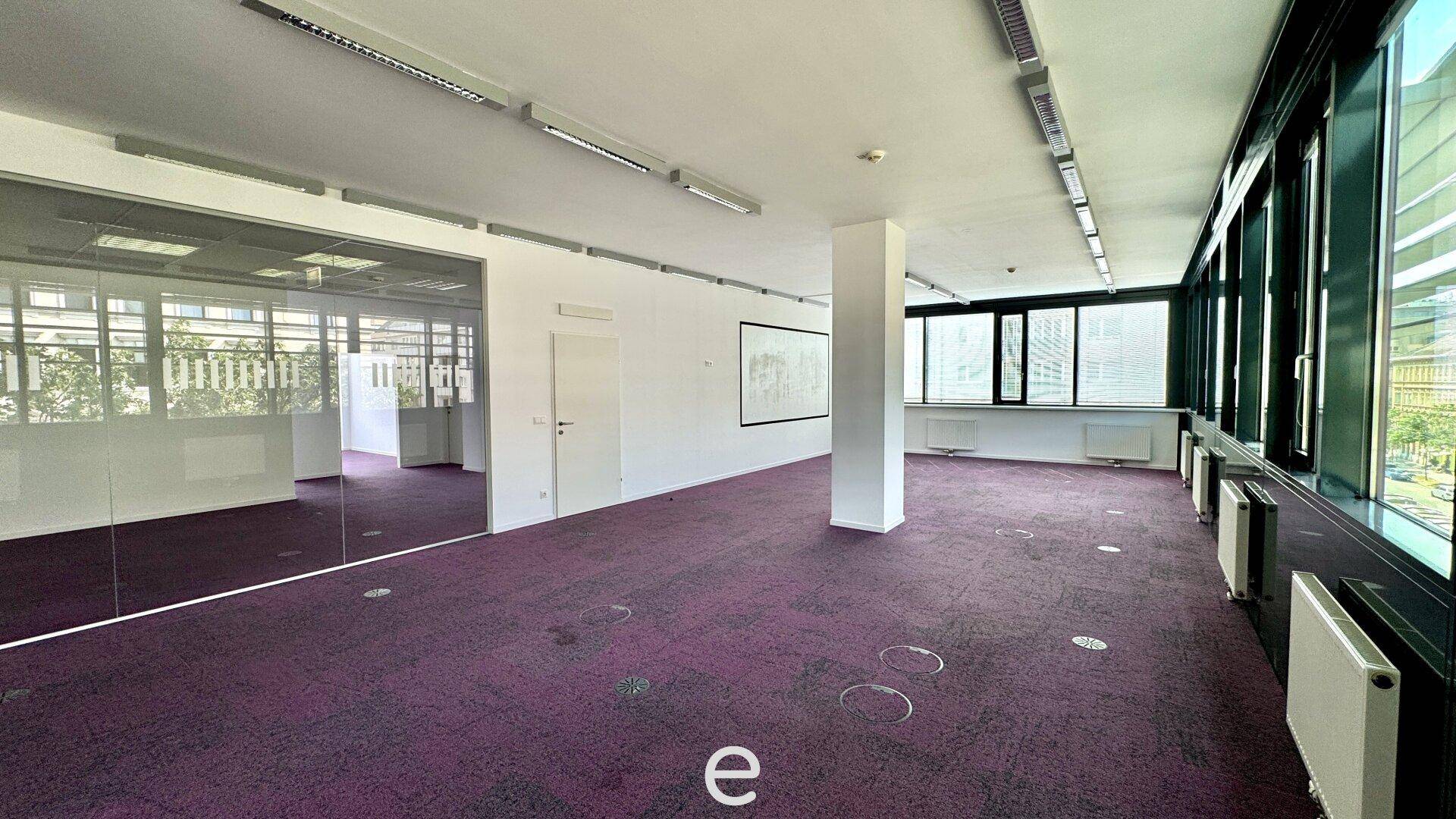 Großes, leeres Büro mit lila Teppichboden, Säulen und einer langen Fensterfront für viel Tageslicht.