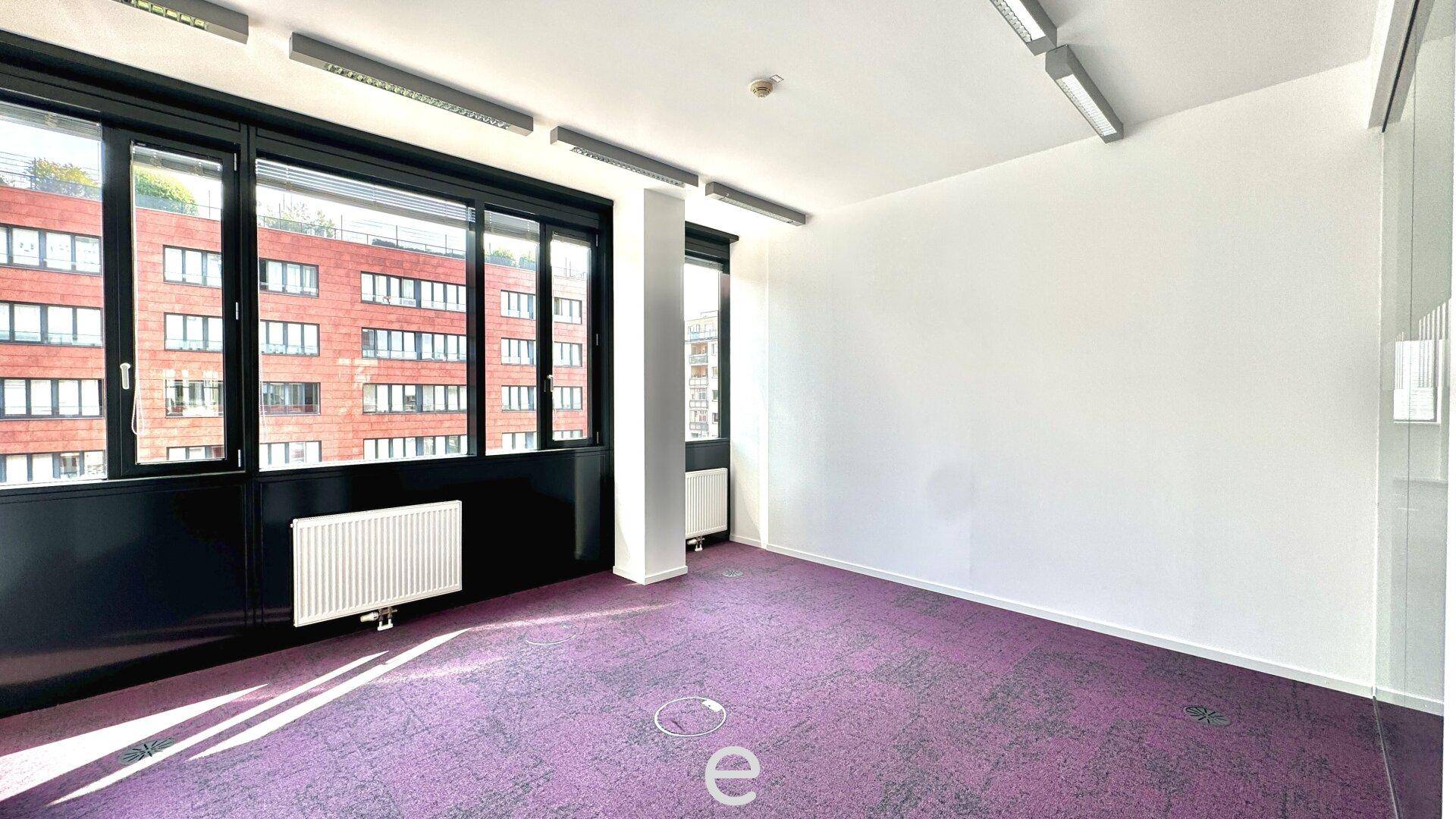 Heller Büroraum mit lila Teppichboden und breiter Fensterfront, die einen Blick nach draußen ermöglicht.