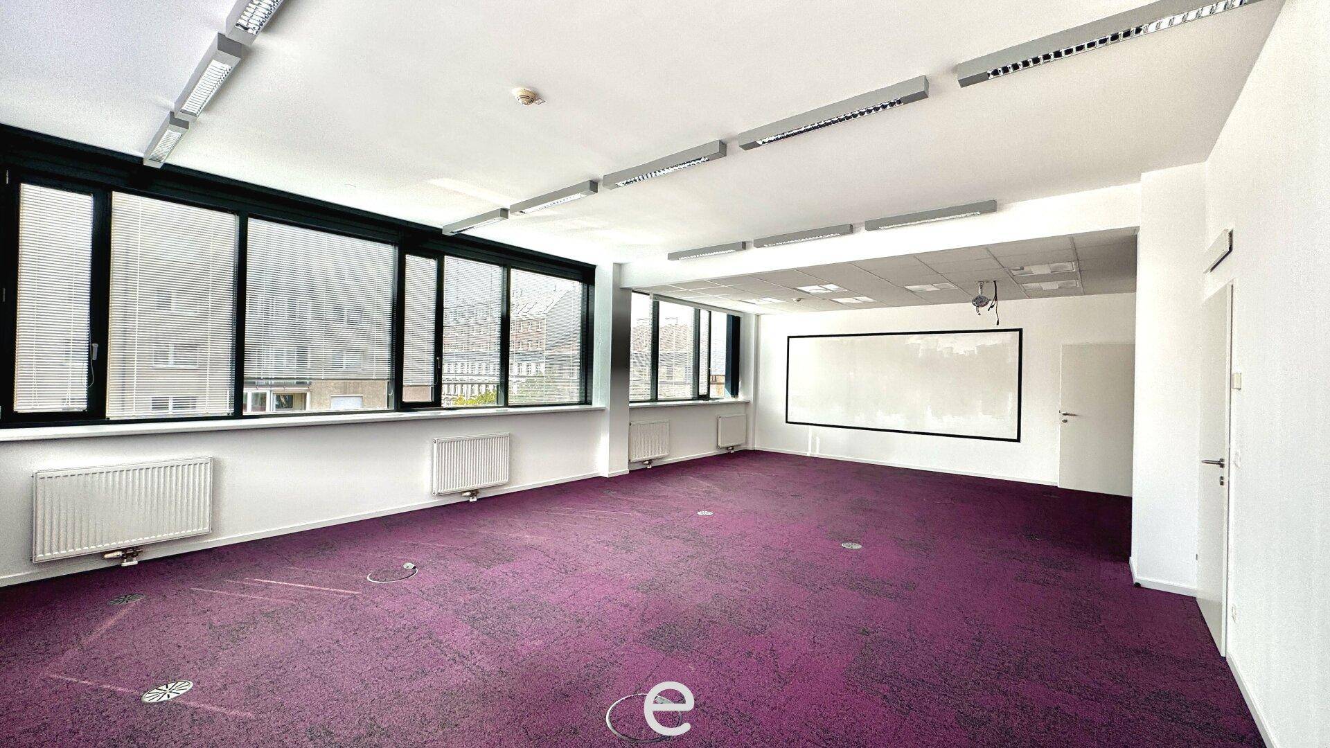 Geräumiges Büro mit lila Teppichboden, großen Fenstern und einer Projektionsfläche an der Wand.