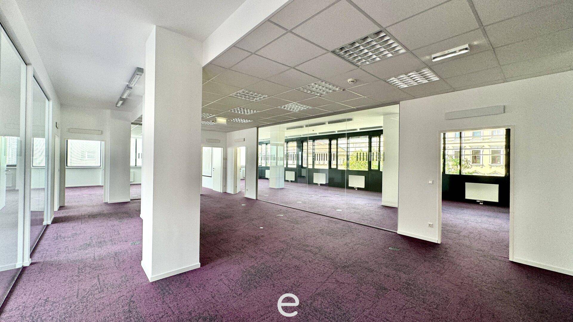 Weitläufiges Büro mit lila Teppichboden, mehreren Säulen und Glaswänden, die den Raum strukturieren.
