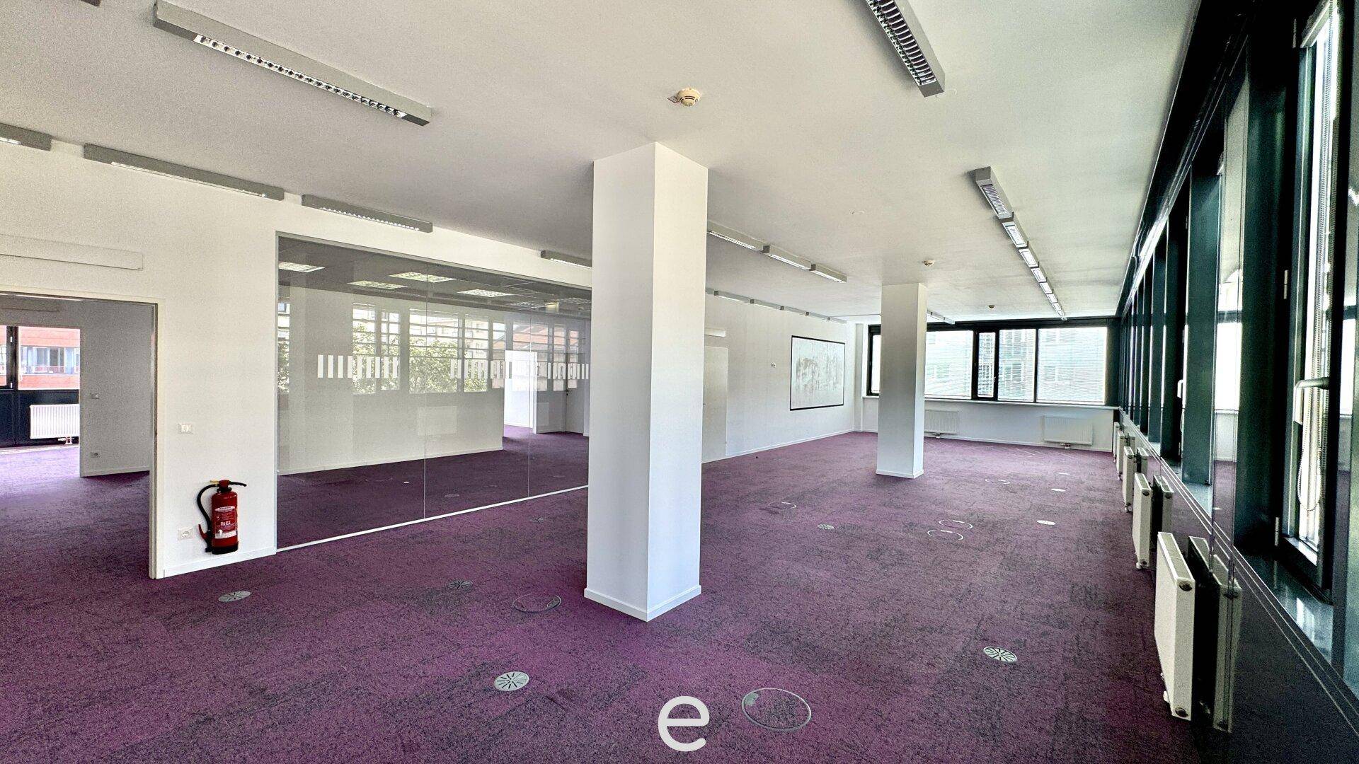 Weitläufiges Büro mit lila Teppichboden, Säulen und einer langen Fensterfront, die viel Licht spendet.