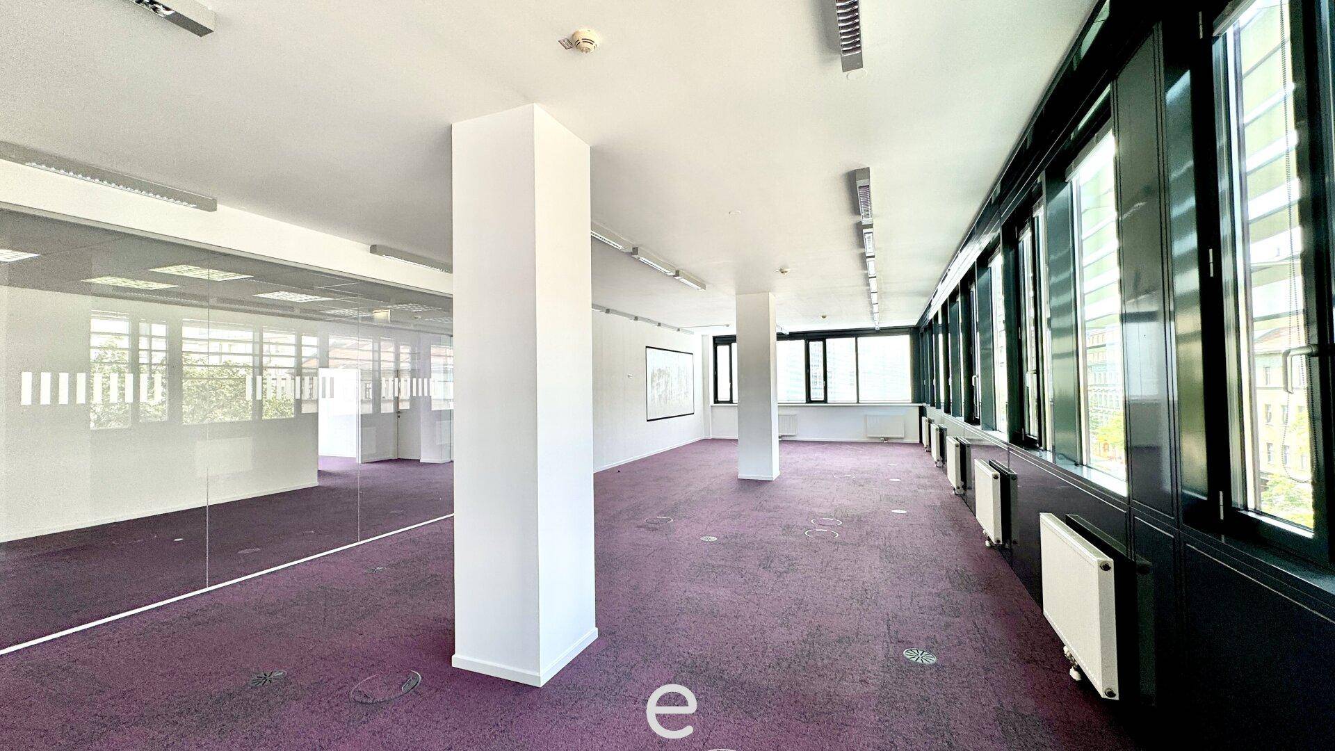Großes, leeres Büro mit lila Teppichboden, Säulen und einer durchgehenden Fensterfront.