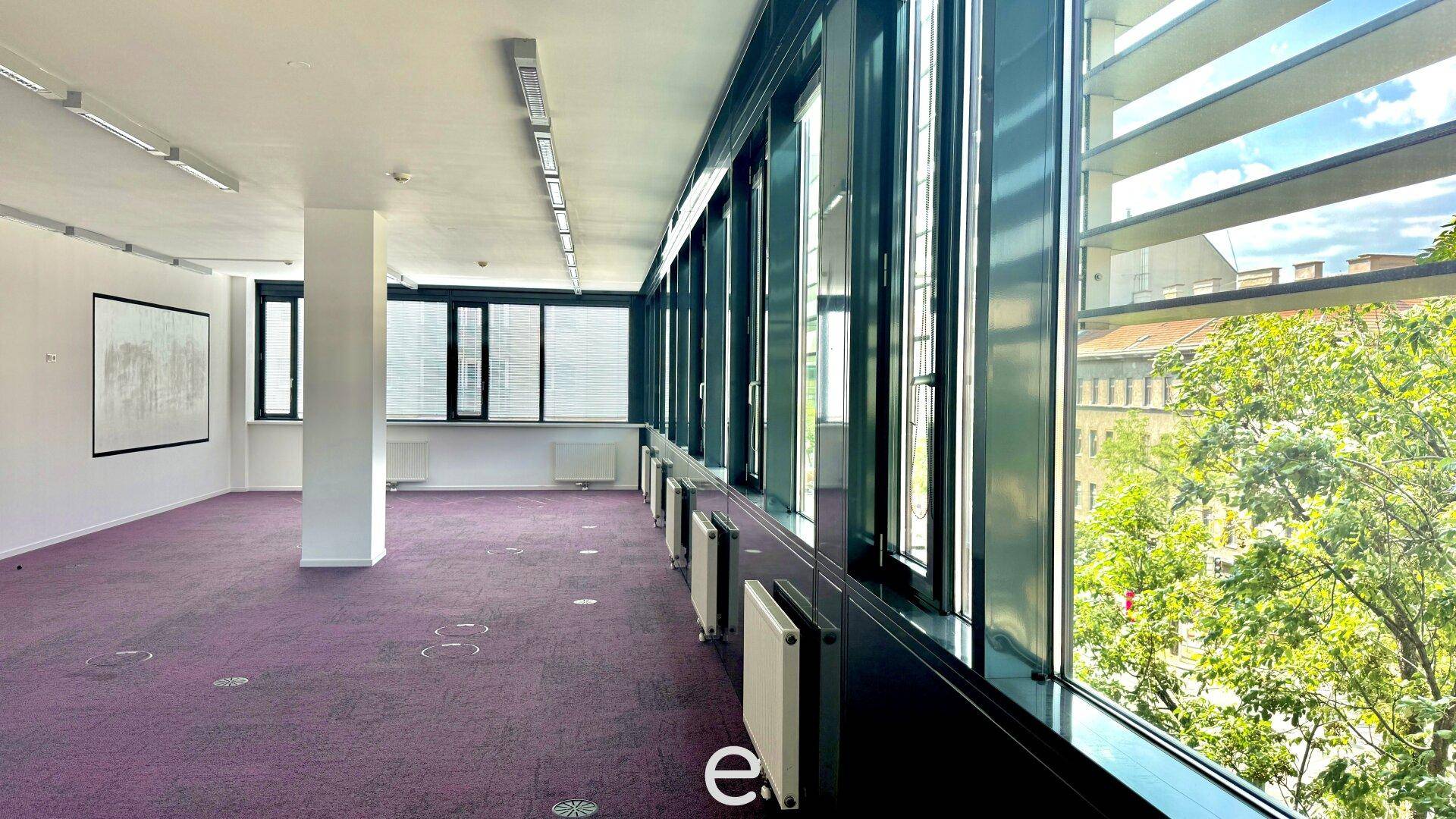 Heller Büroraum mit lila Teppichboden und breiter Fensterfront, die einen Blick auf Bäume bietet.