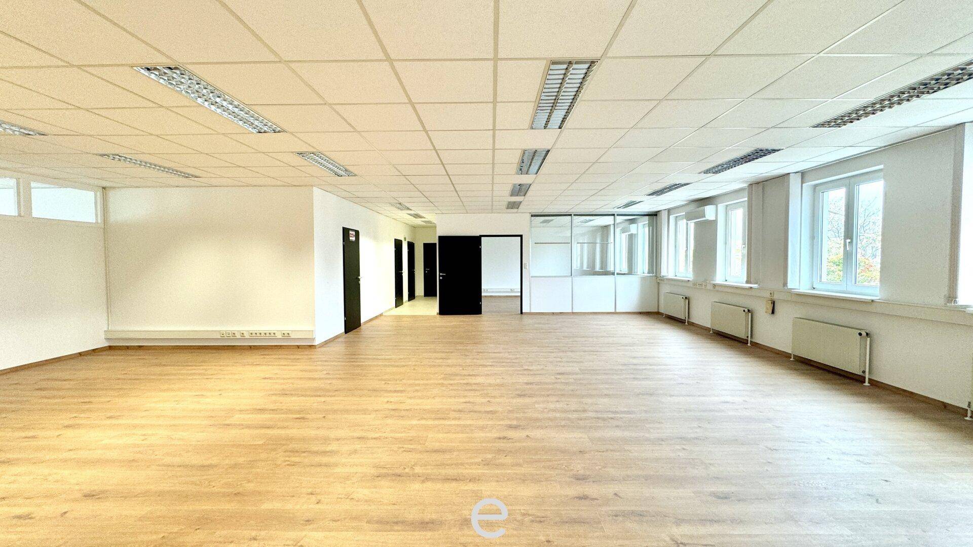 Großer, heller Büroraum mit Holzfußboden, abgehängter Decke und vielen Fenstern.
