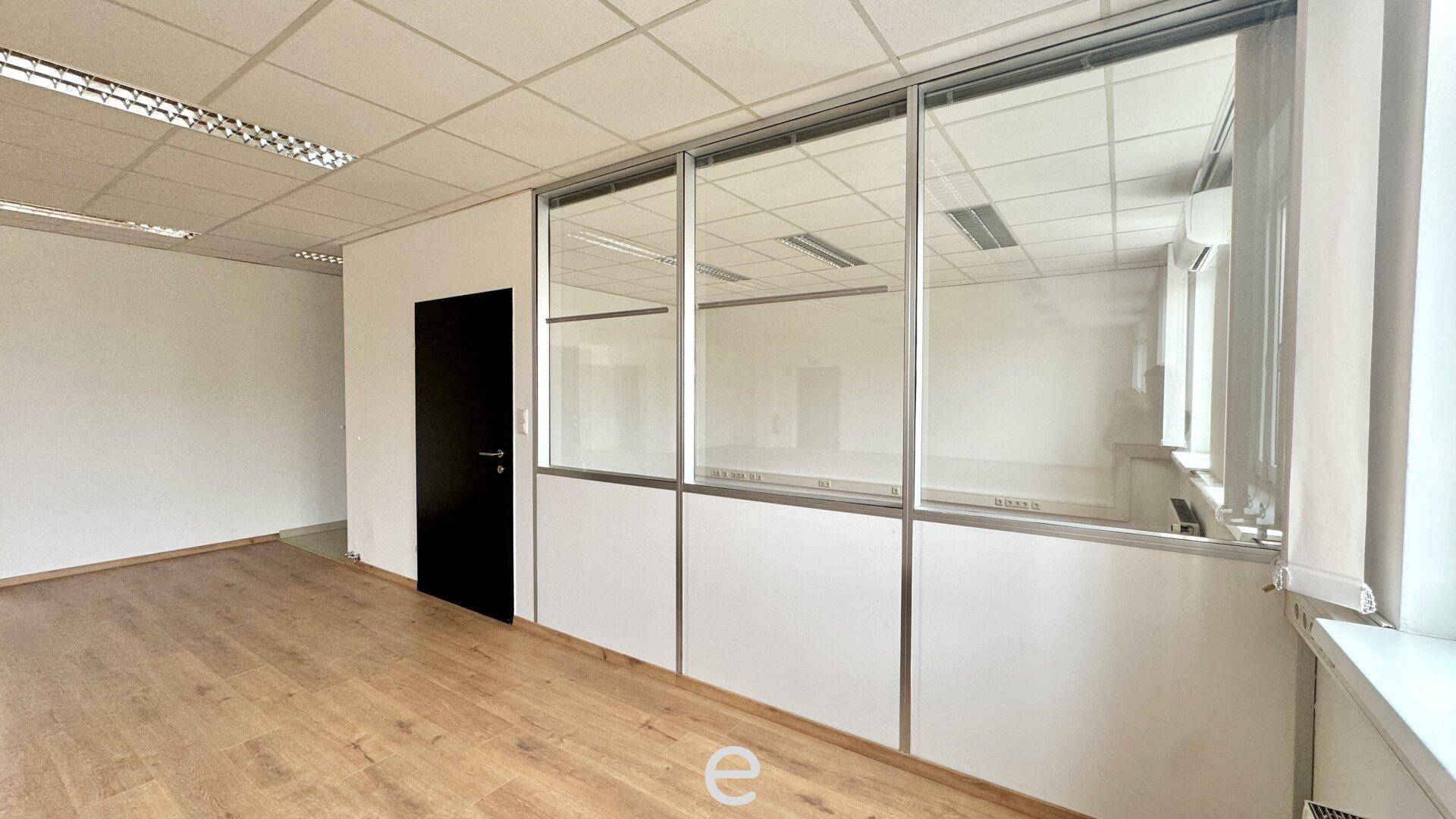 Büroraum mit Holzfußboden, weißer Wand und einer Glaswand, die einen weiteren Bereich abtrennt.