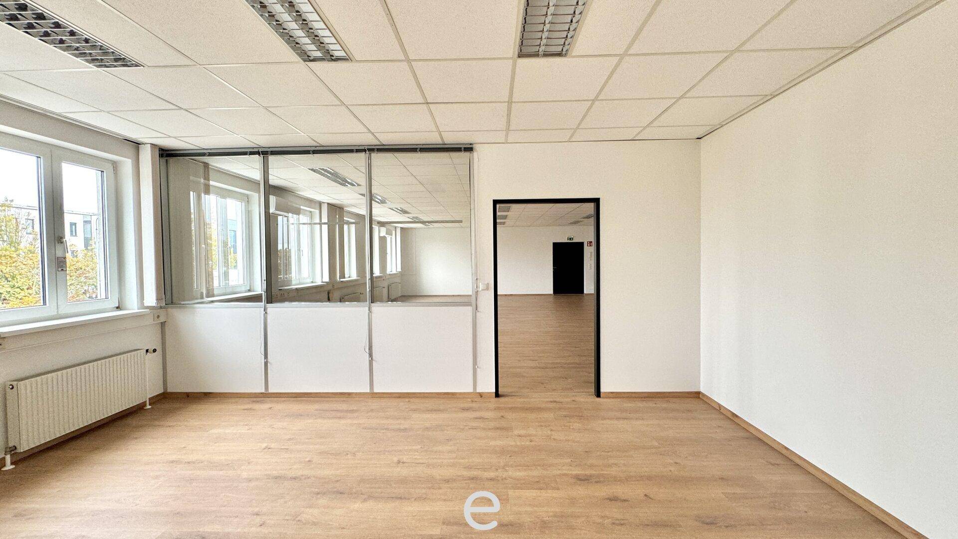 Heller Büroraum mit Holzfußboden, Glaswänden und Blick auf weitere Bürobereiche.