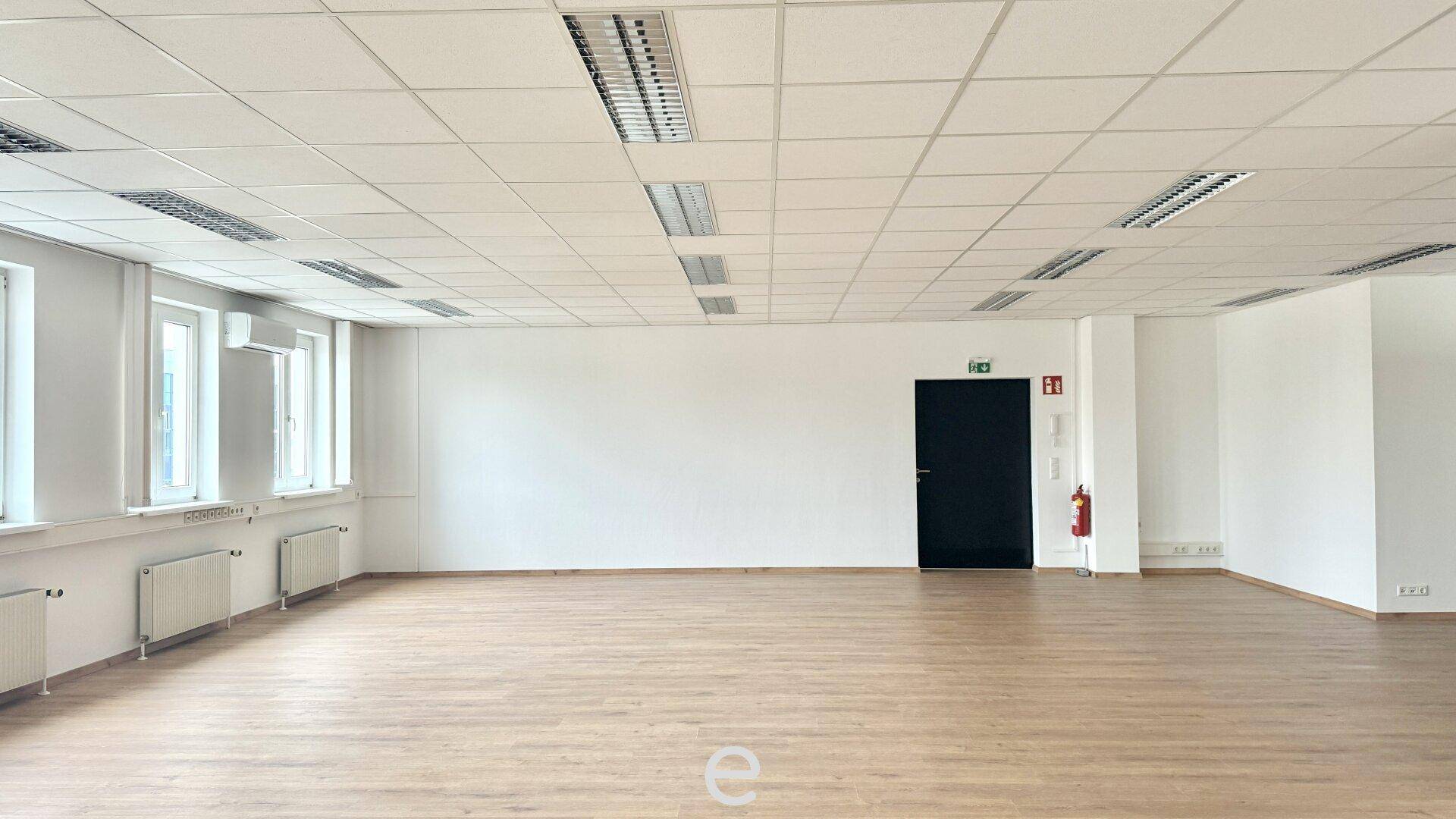 Großer, leerer Büroraum mit Holzfußboden, weißen Wänden und vielen Fenstern.
