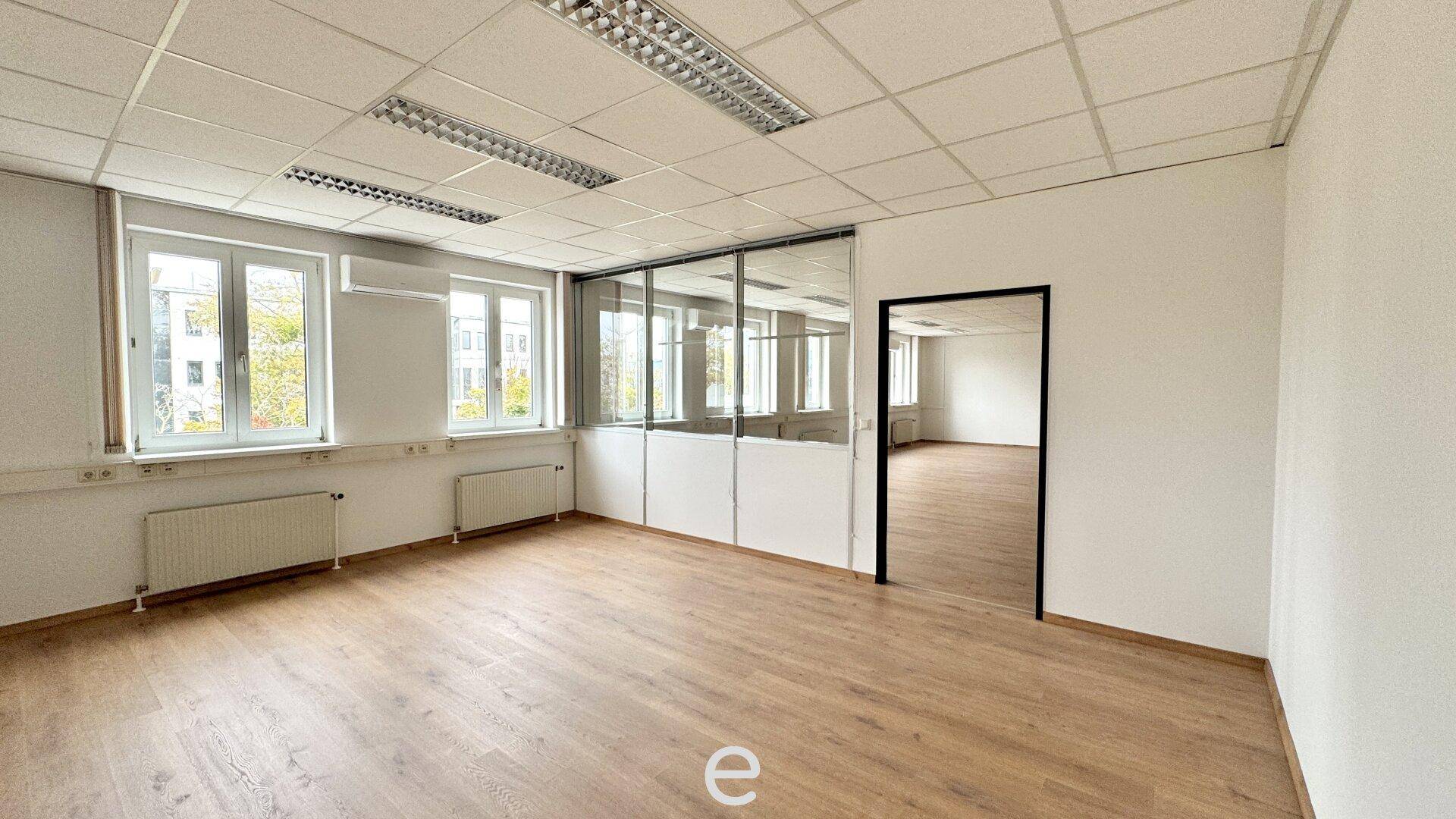 Heller Büroraum mit Holzfußboden, Glaswänden und Blick in einen weiteren Bürobereich.