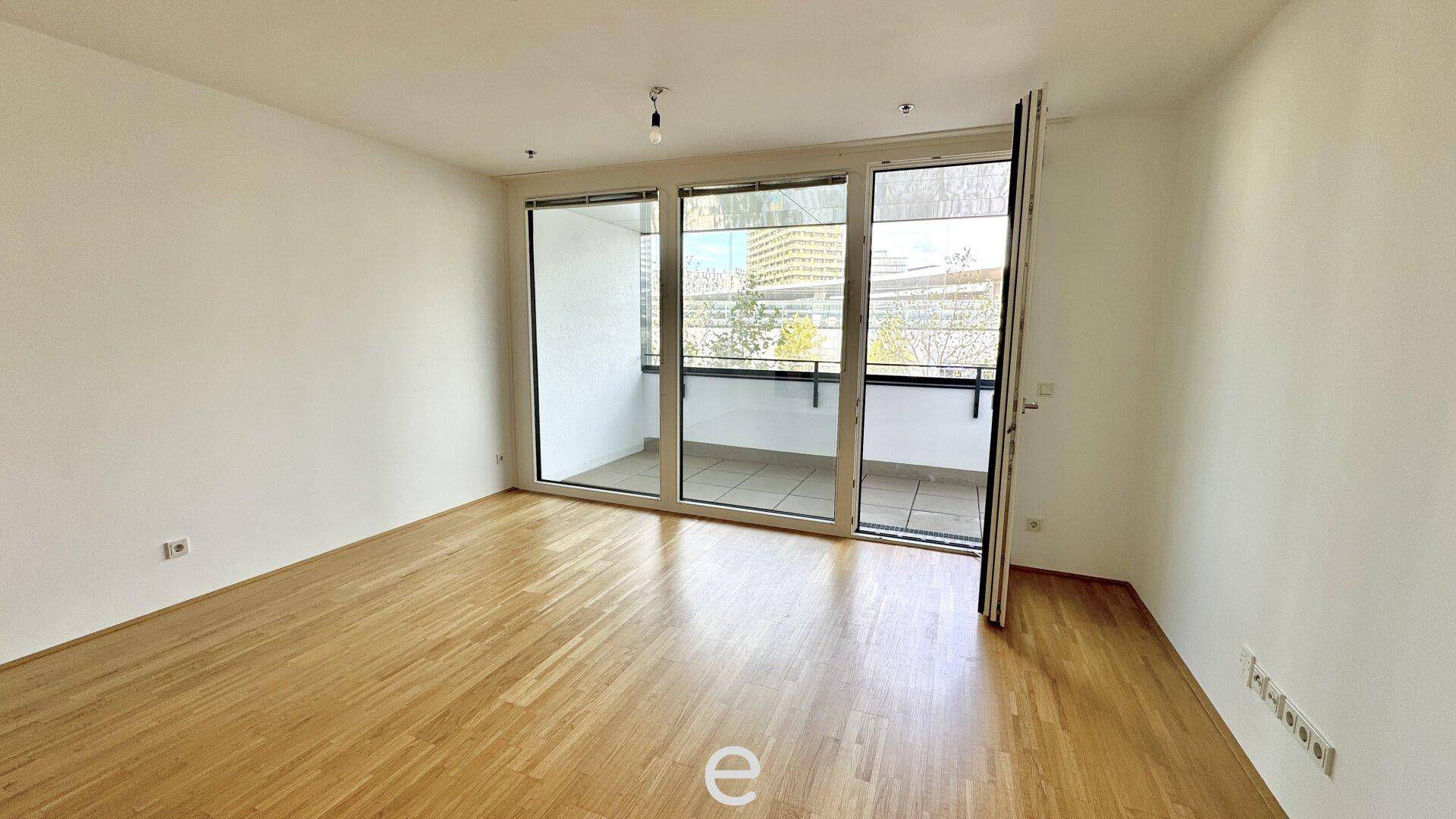 Leerer Wohnraum mit Holzboden und großen Glasfronten, die zu einem Balkon oder einer Loggia führen.