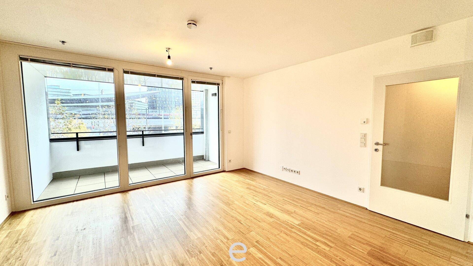 Geräumiger Wohnraum mit Holzboden und großen Glasfronten, die Zugang zu einem Außenbereich bieten.