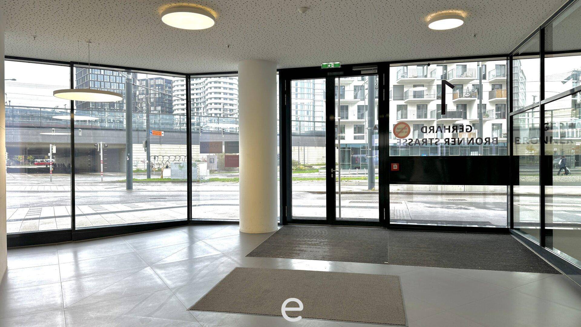 Heller Eingangsbereich mit großen Glasfronten, einer Säule und modernen Deckenleuchten.