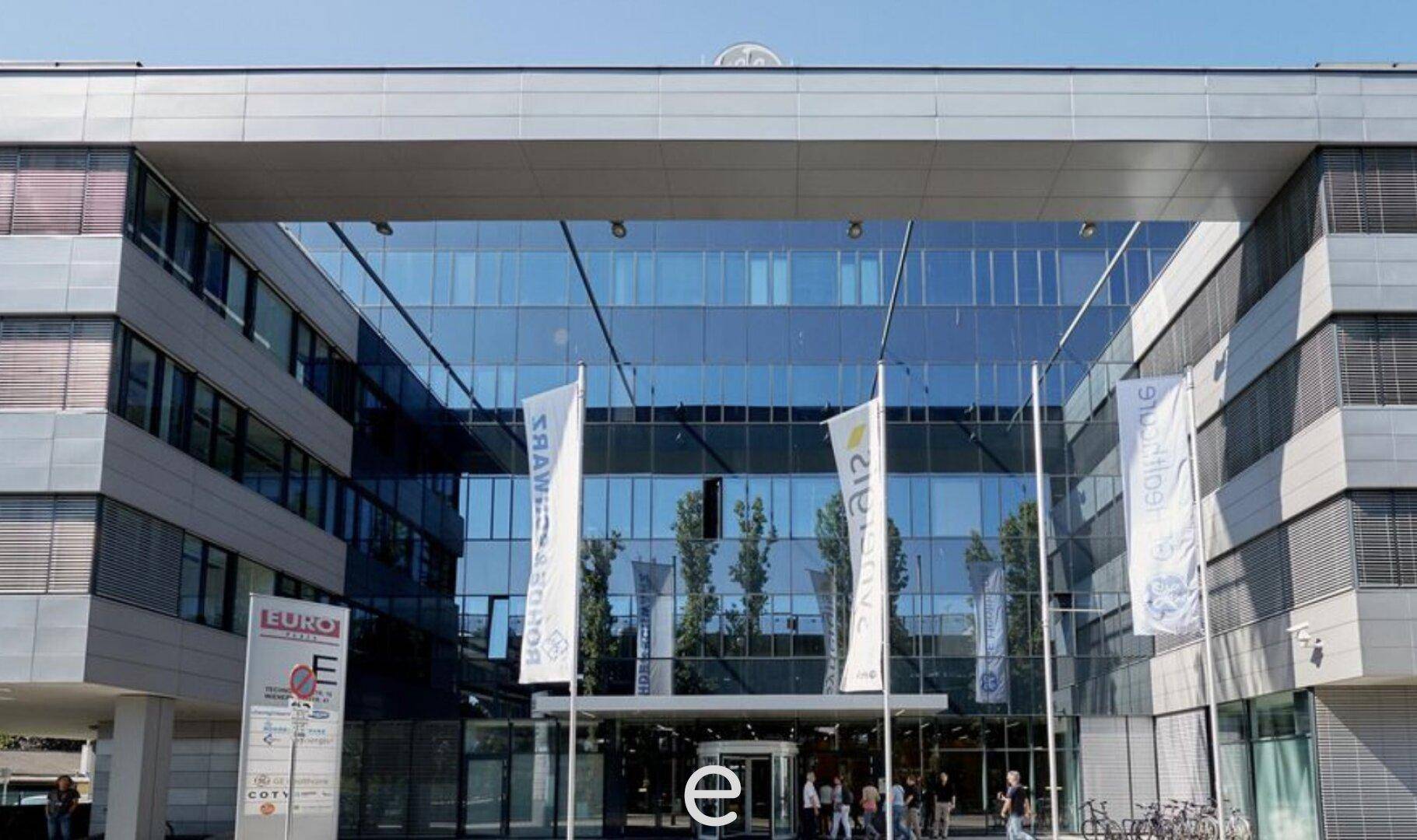 Haupteingang eines modernen Bürokomplexes mit großer Glasfront und Firmenflaggen.
