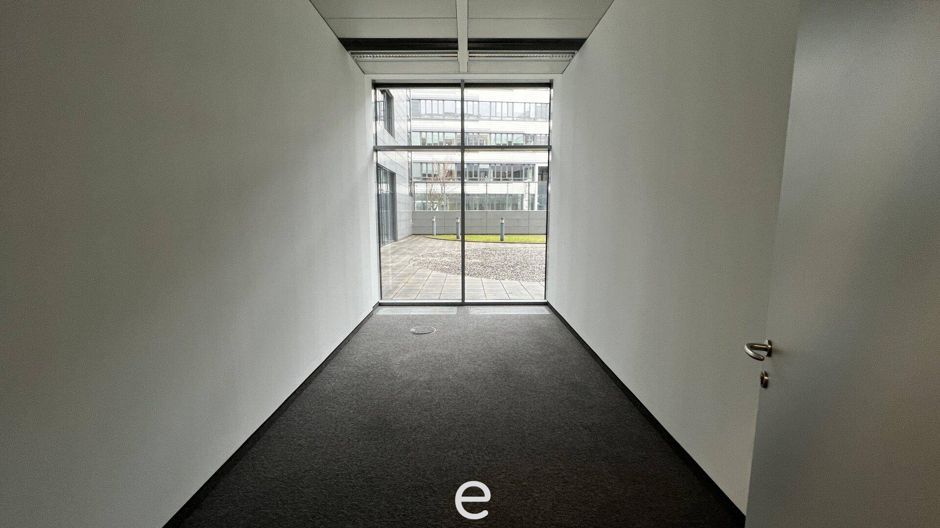 Kleiner Büroraum mit Teppichboden und einer großen Fensterfront, die den Blick nach draußen freigibt.