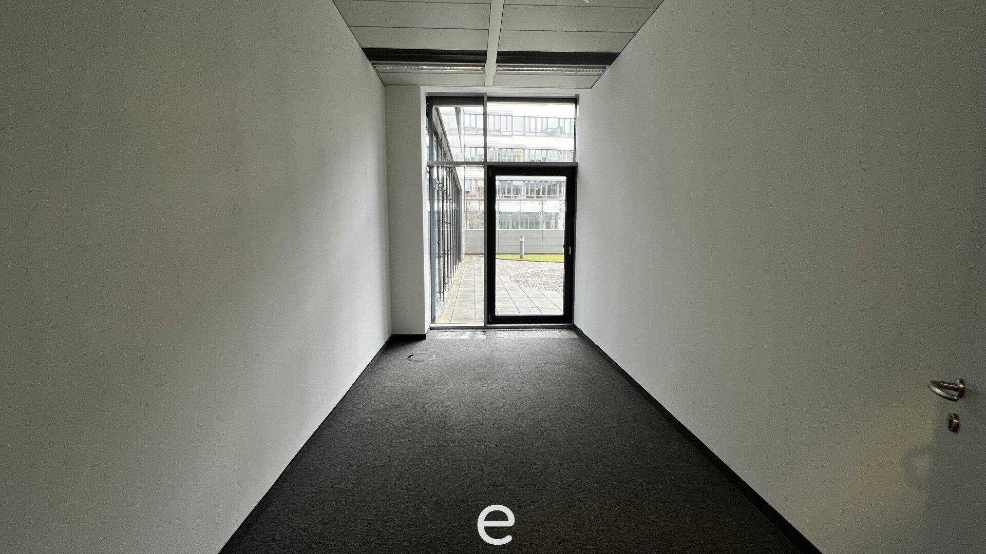 Schmaler Büroraum mit Teppichboden und einer Glastür, die zu einem Außenbereich führt.