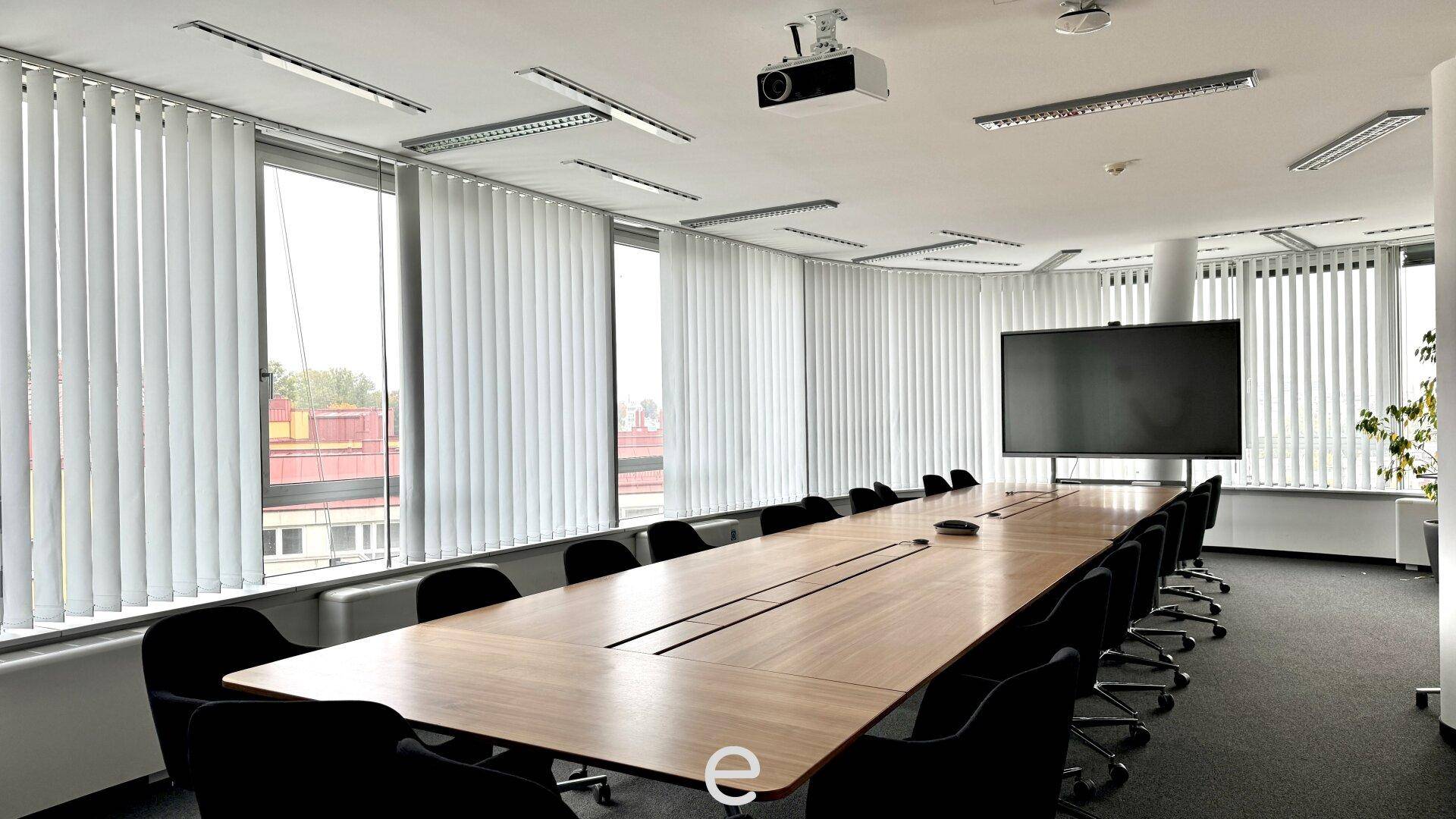 Großer Konferenzraum mit langem Holztisch, vielen Stühlen und vertikalen Jalousien an den Fenstern.
