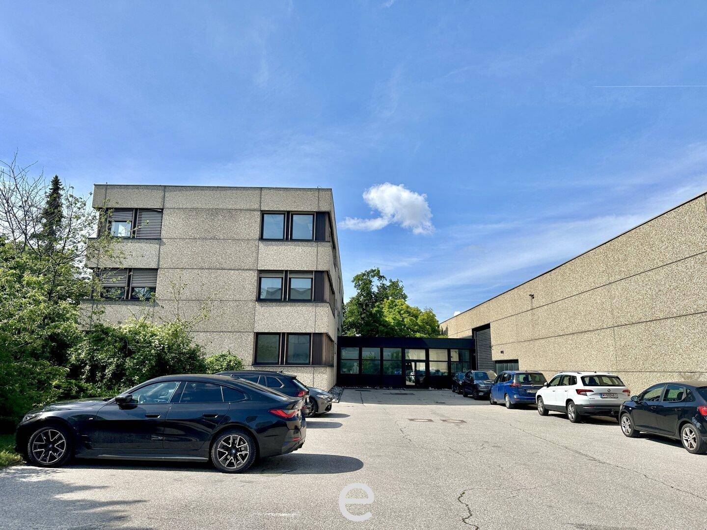 Ansicht des Bürogebäudes und der Lagerhalle mit Parkplätzen unter blauem Himmel.