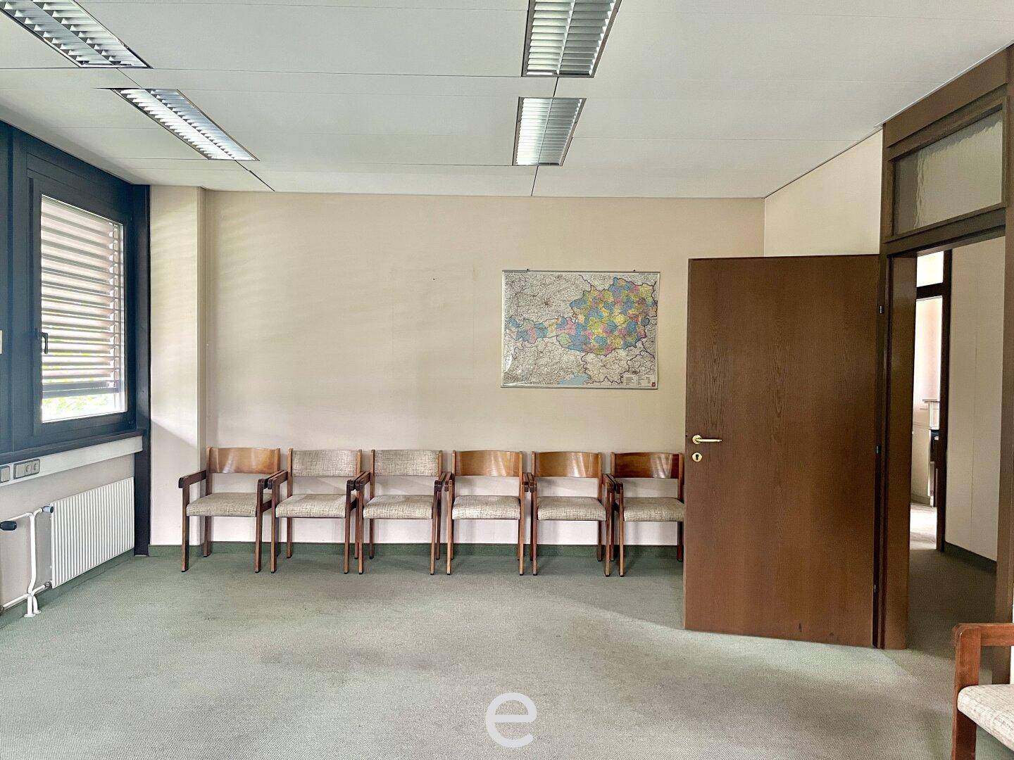 Einfacher Wartebereich oder Besprechungsraum mit Teppichboden, Fenster und einer Reihe von Stühlen.