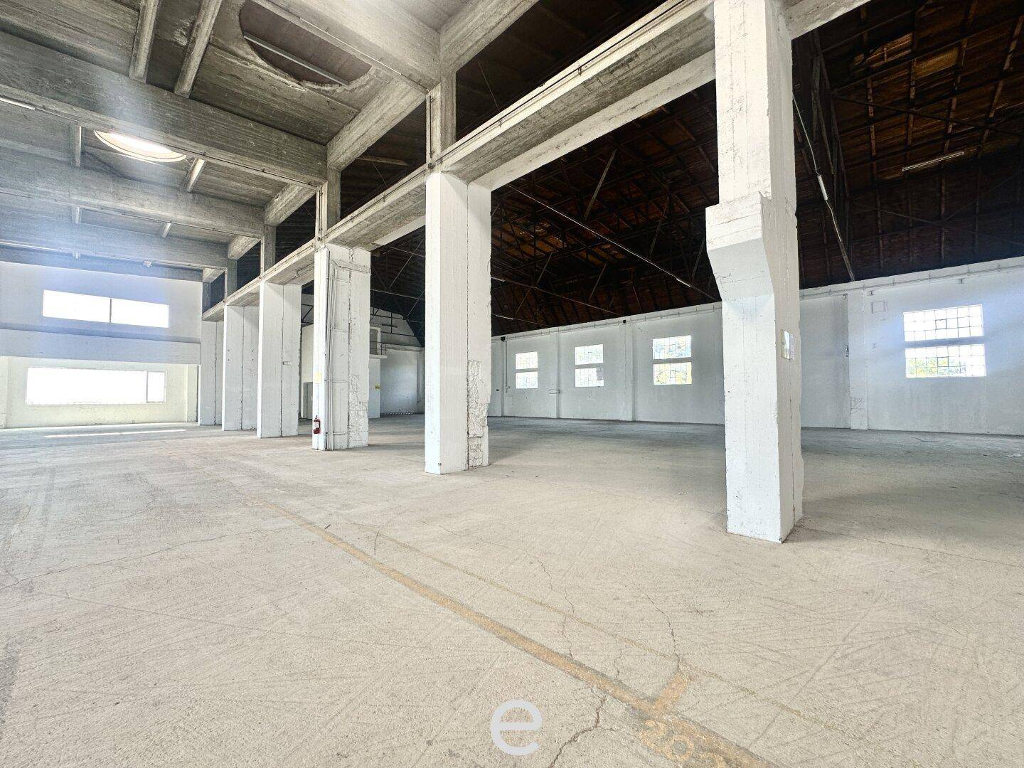 Weitläufige, leere Lagerhalle mit Betonboden, hohen Decken und sichtbarer Holz-Dachkonstruktion.