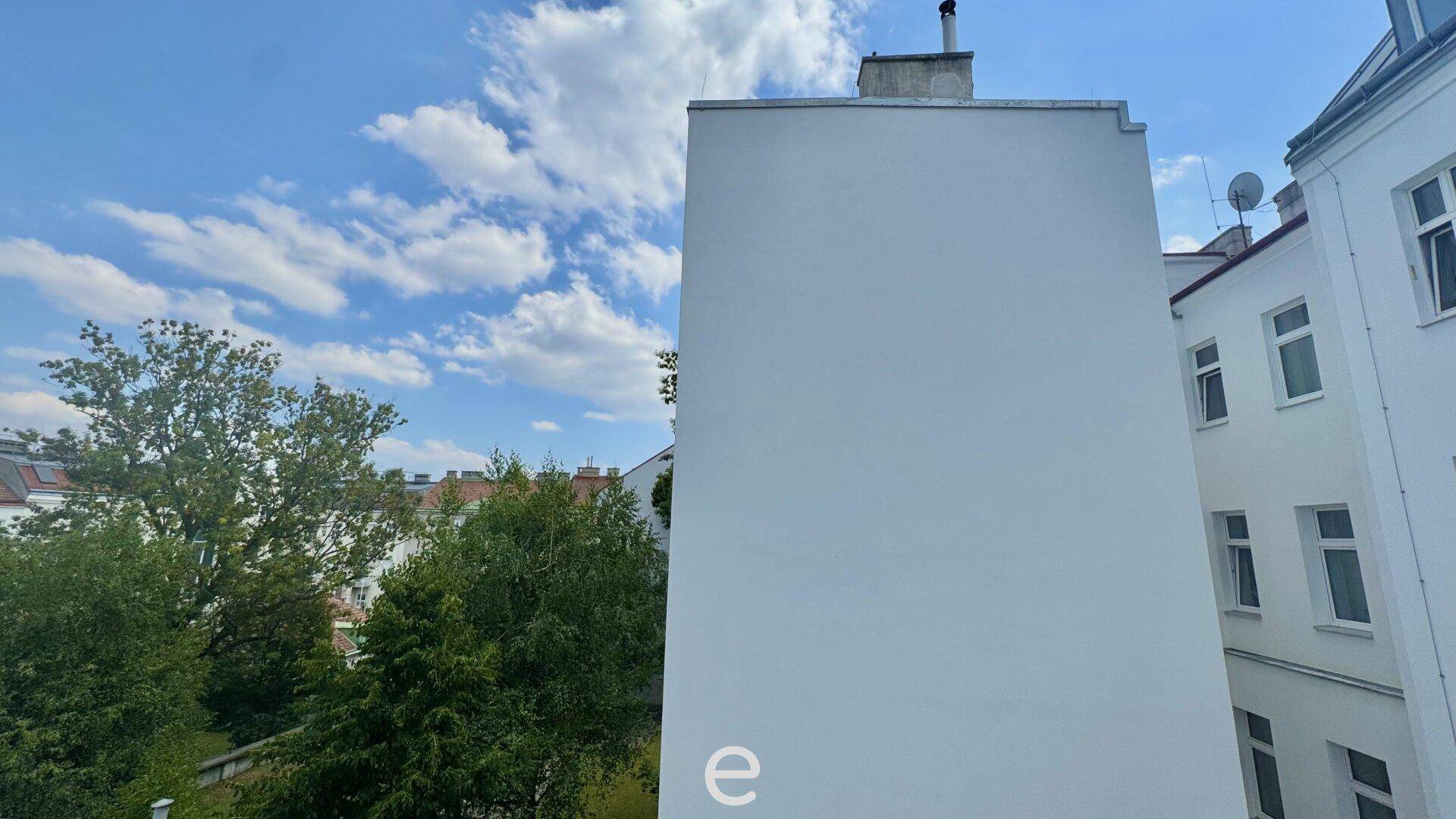 Blick auf die Seitenfassade eines weißen Gebäudes unter einem blauen Himmel mit Wolken und Bäumen.