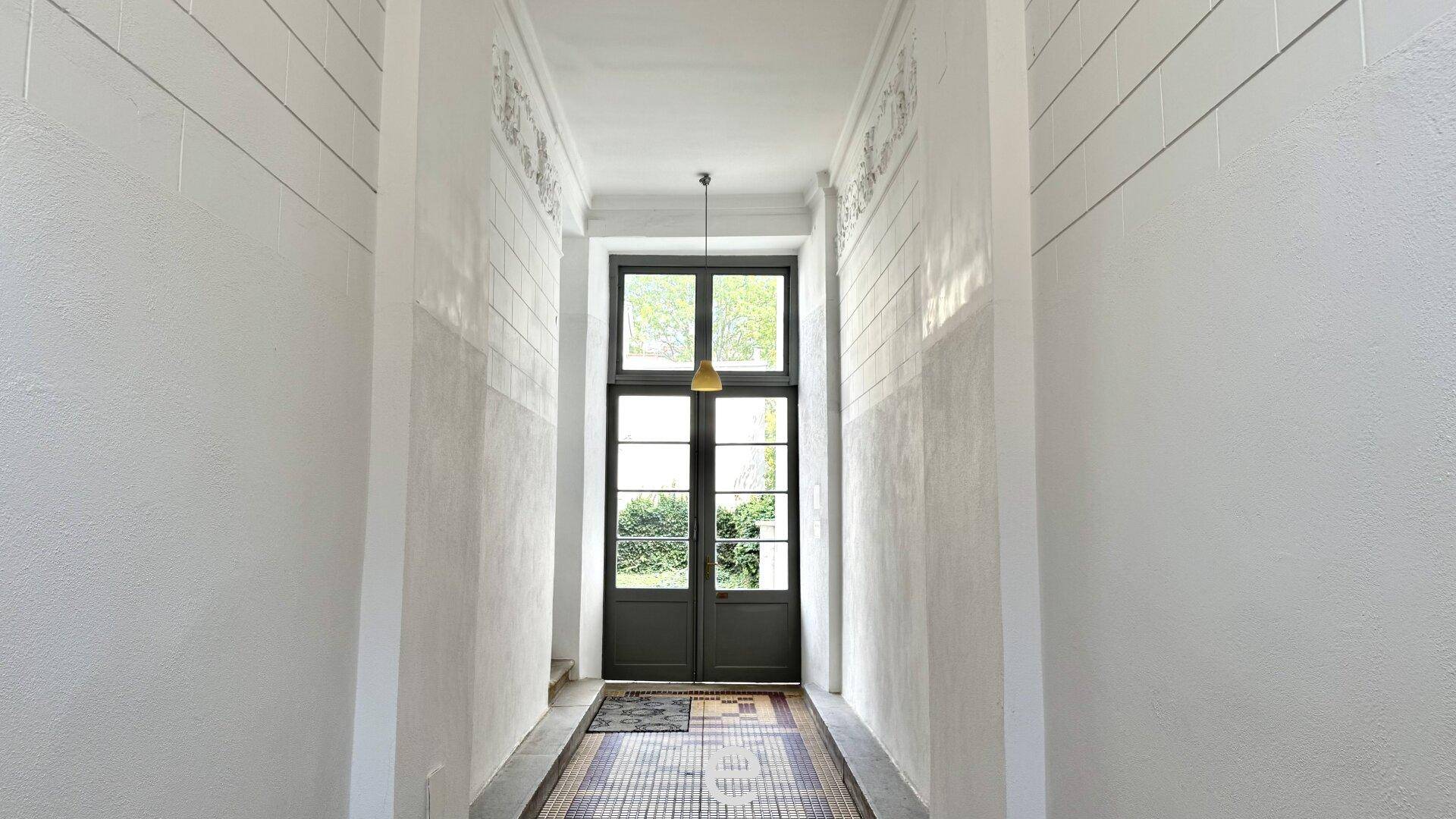 Heller Flur mit hohen Decken, Stuckverzierungen und einer großen Glastür zum Außenbereich.