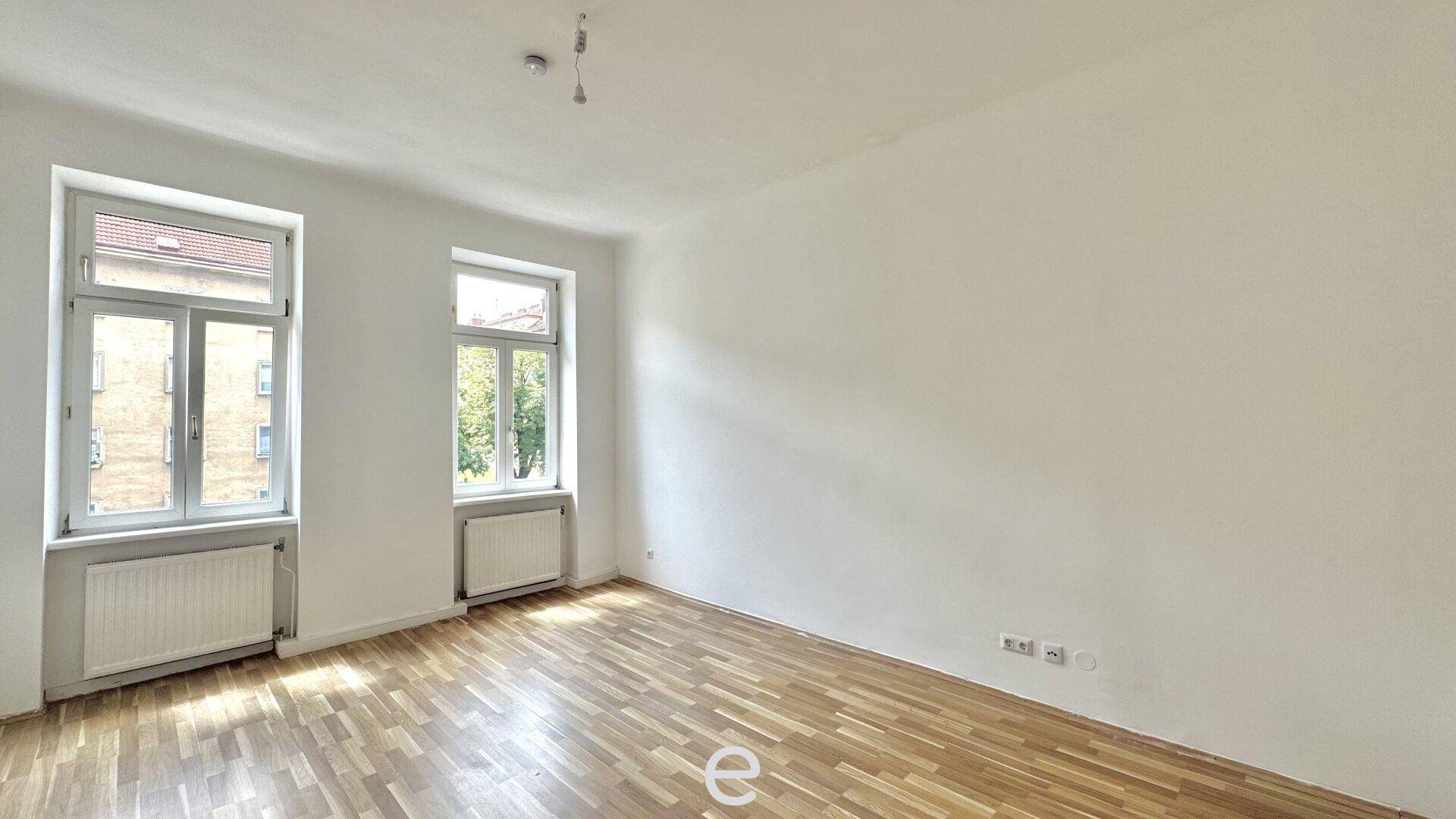 Geräumiger, leerer Wohnraum mit Holzboden und viel Tageslicht durch zwei Fenster.