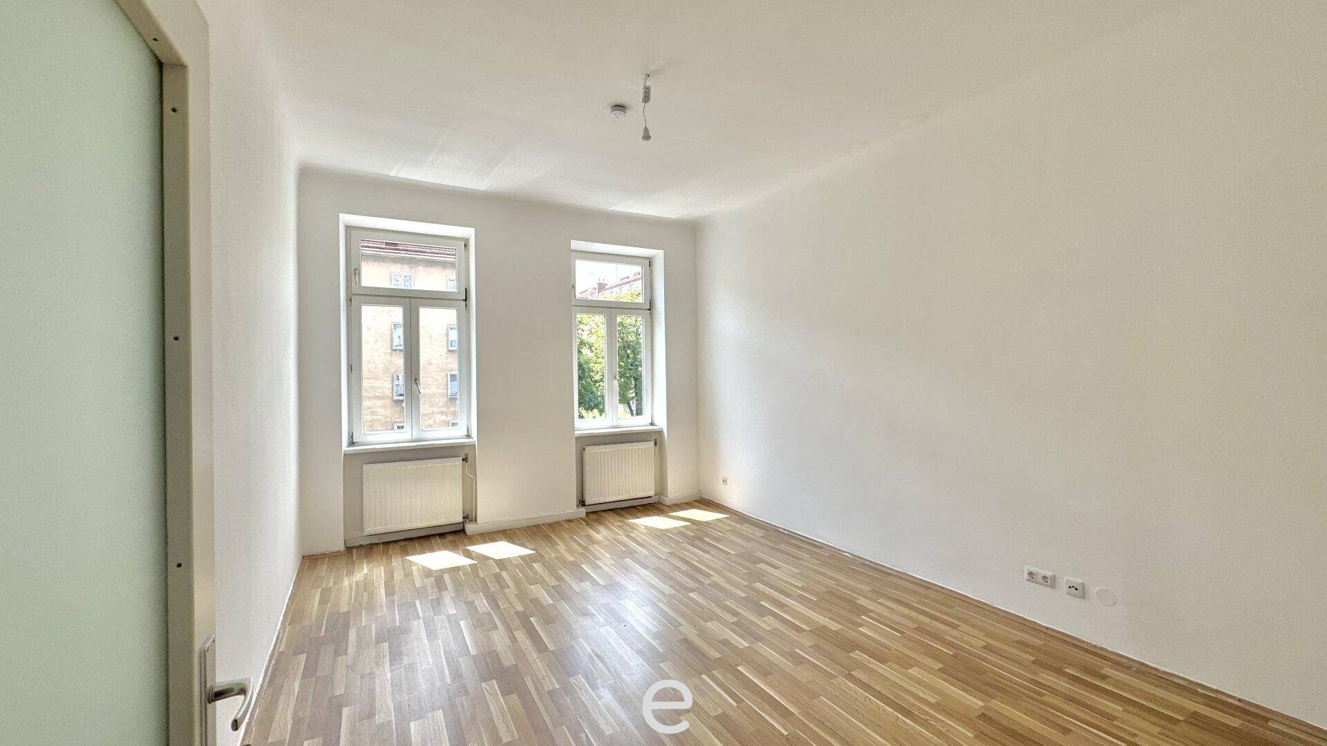 Heller, leerer Wohnraum mit Parkettboden und zwei großen Fenstern zur Straße.