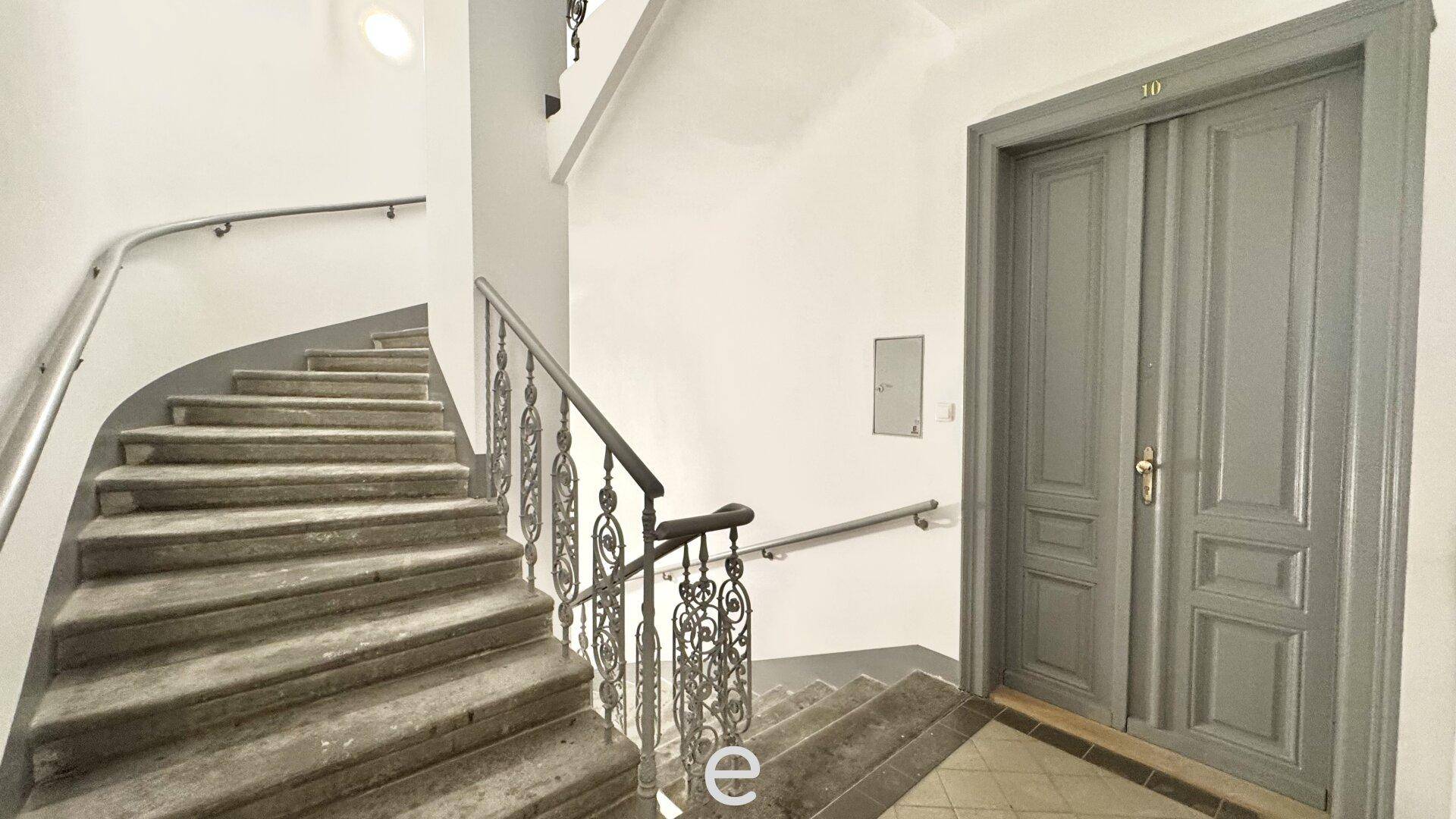 Elegantes Treppenhaus mit geschwungener Steintreppe, kunstvollem Geländer und einer großen Doppeltür im Altbau-Stil.