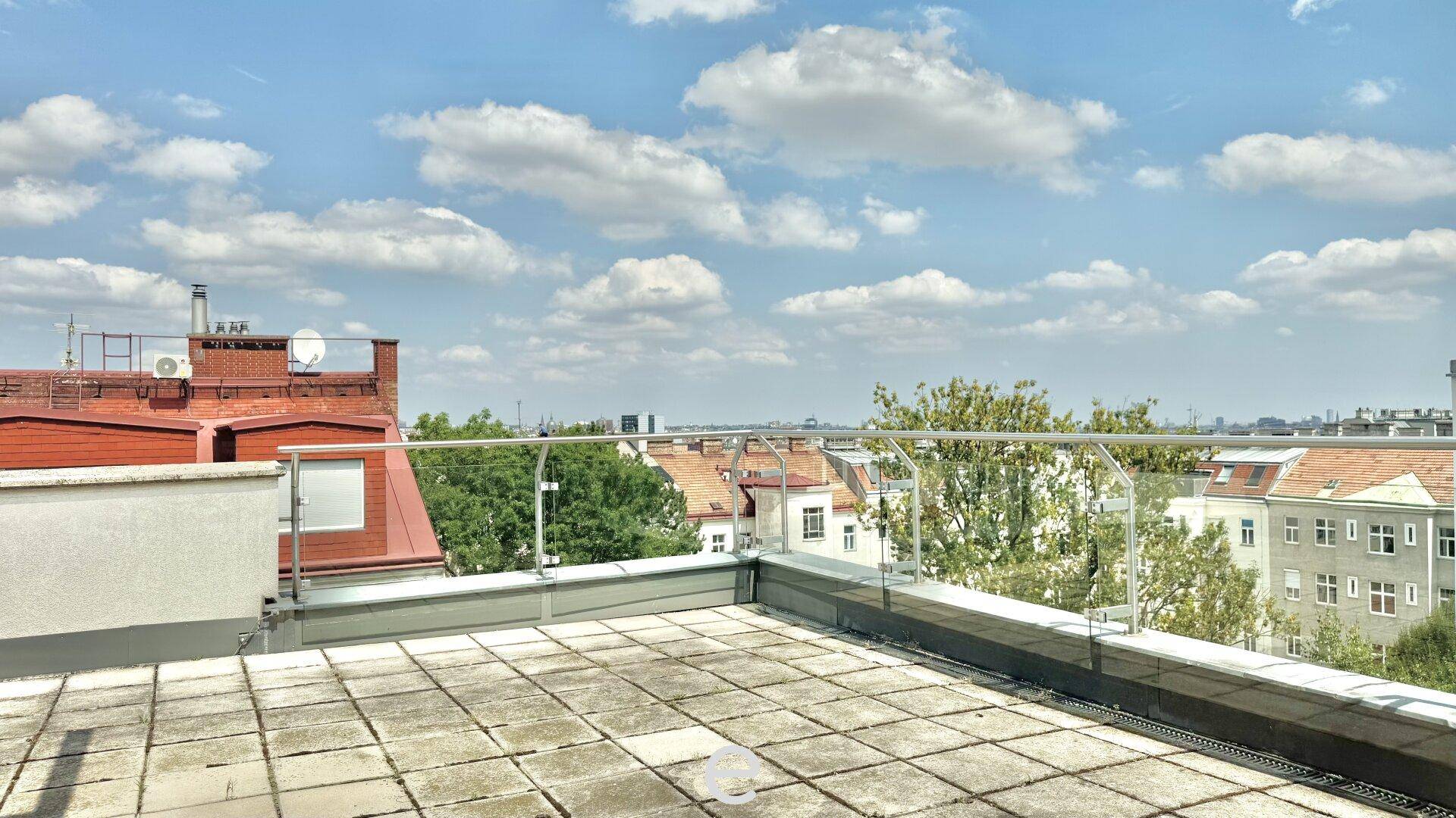 Geräumige Dachterrasse mit Fliesenboden, Glasgeländer und weitem Blick über die Stadt.