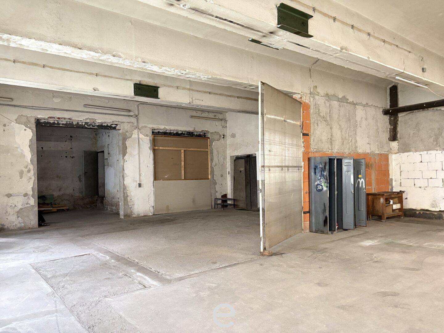 Weitläufiger Industrieraum mit Betonboden, hohen Decken, einem großen Metalltor und alten Spinden.