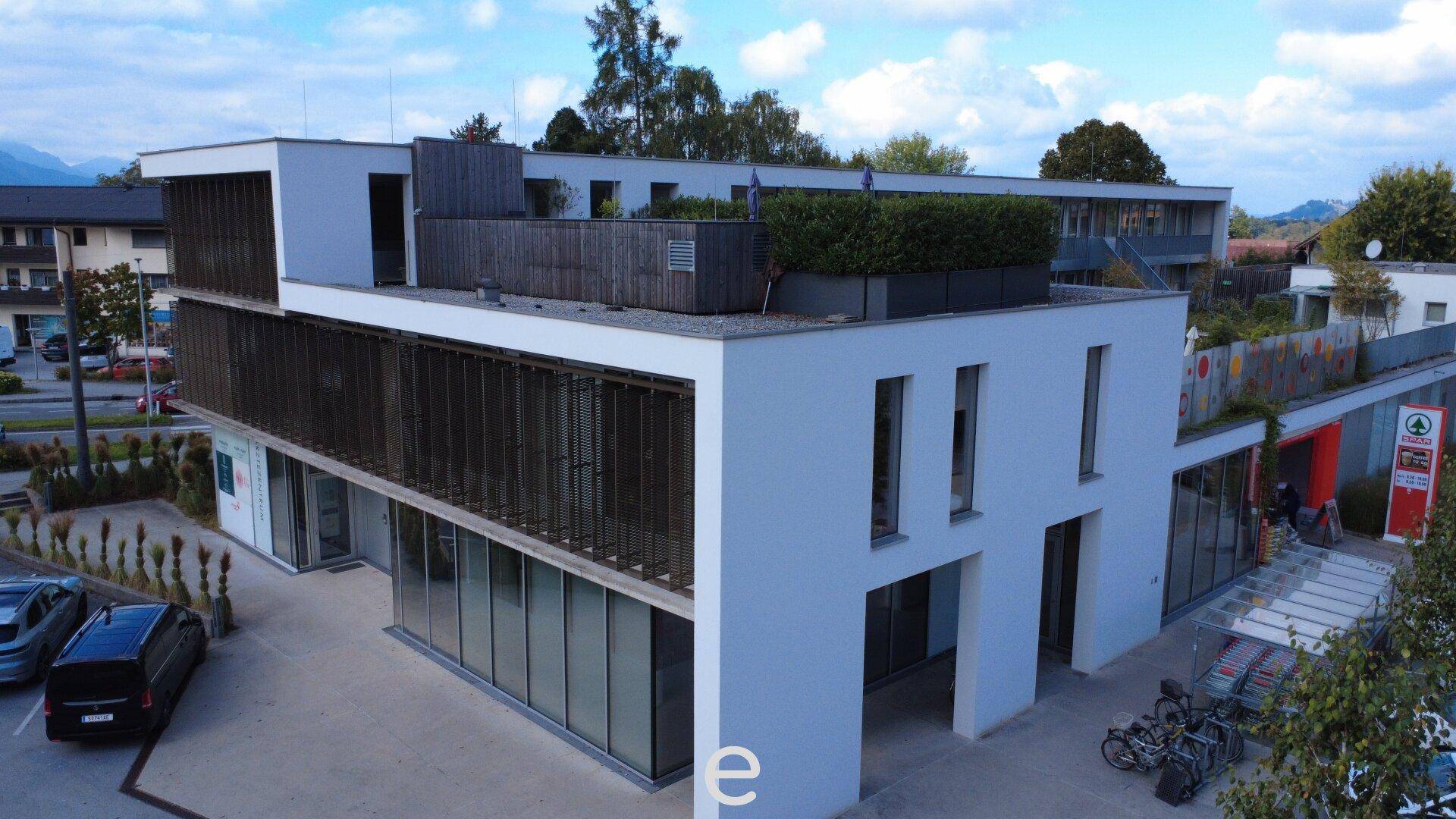 Das moderne Gebäude des Ärztezentrums mit Parkplätzen und einer begrünten Dachterrasse, umgeben von Bäumen.