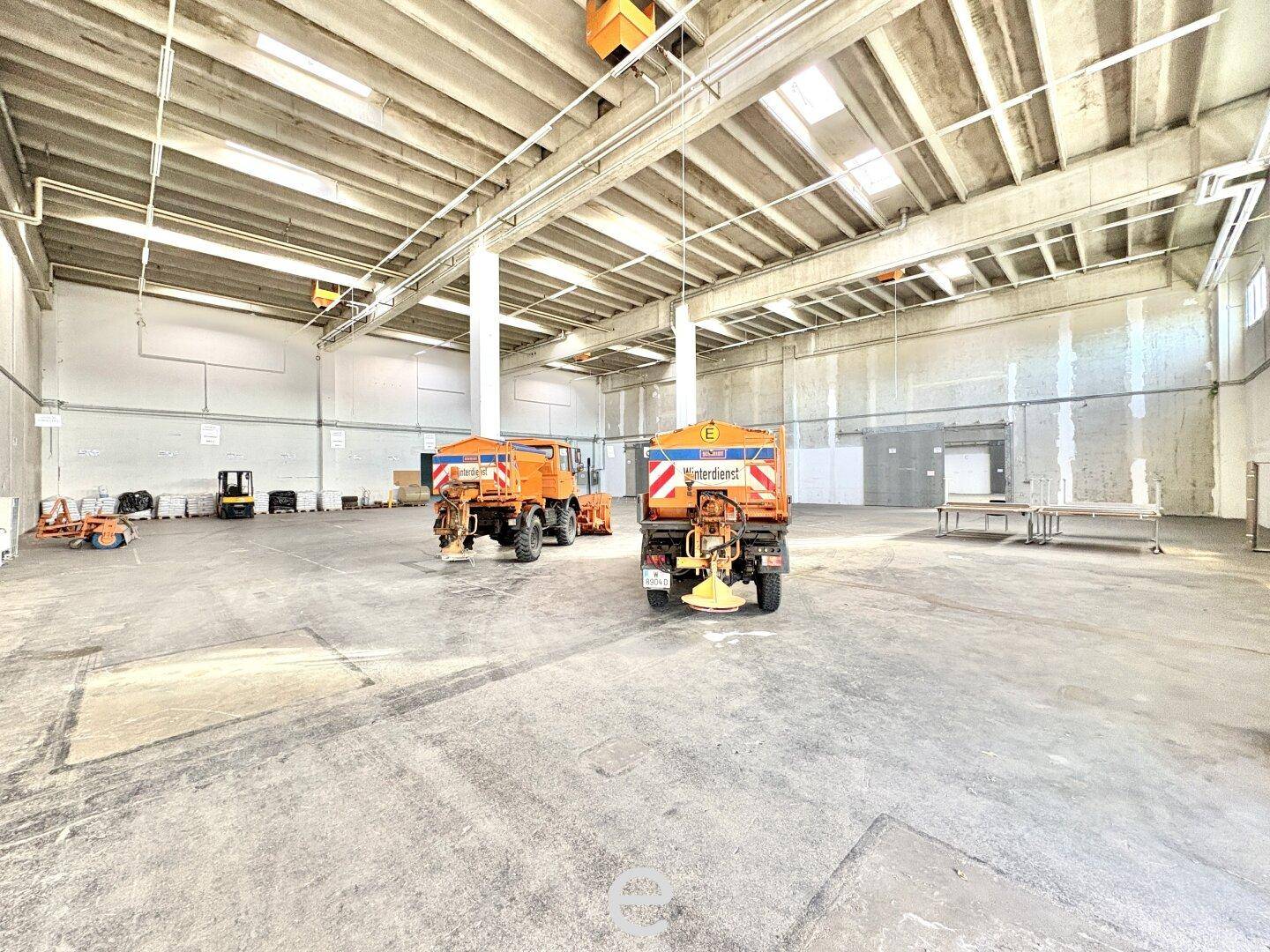 Weitläufige Industriehalle mit zwei großen orangefarbenen Schneepflügen, Betonboden und hohen Decken.