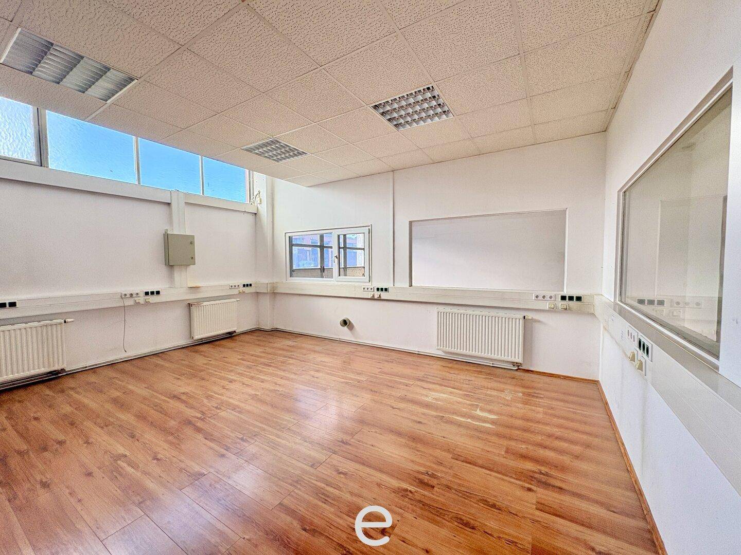 Heller Büroraum mit Laminatboden, abgehängter Decke und großen Fenstern, ausgestattet mit Heizkörpern.
