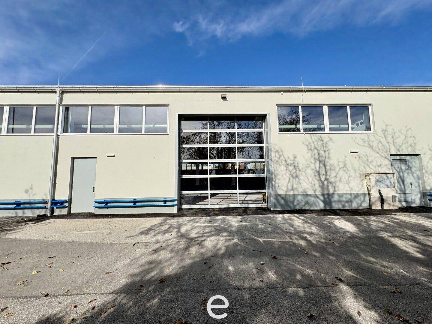 Außenansicht des Gebäudes mit großer Glas-Sektionaltor und mehreren Fenstern unter blauem Himmel.