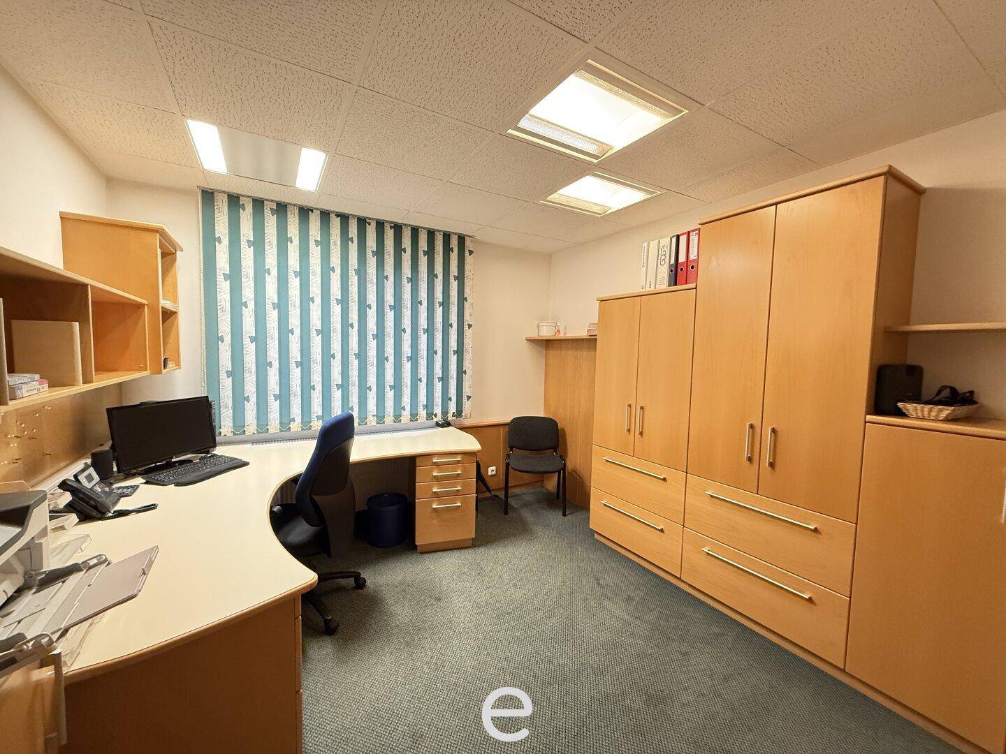 Einzelbüro mit Teppichboden, Holzschränken und einem Schreibtisch am Fenster.