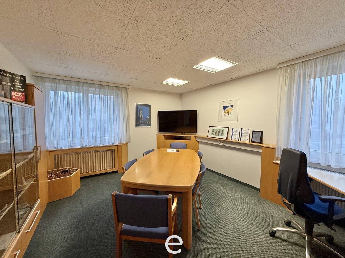 Besprechungsraum mit Teppichboden, großem Holztisch und Fenstern mit Vorhängen.