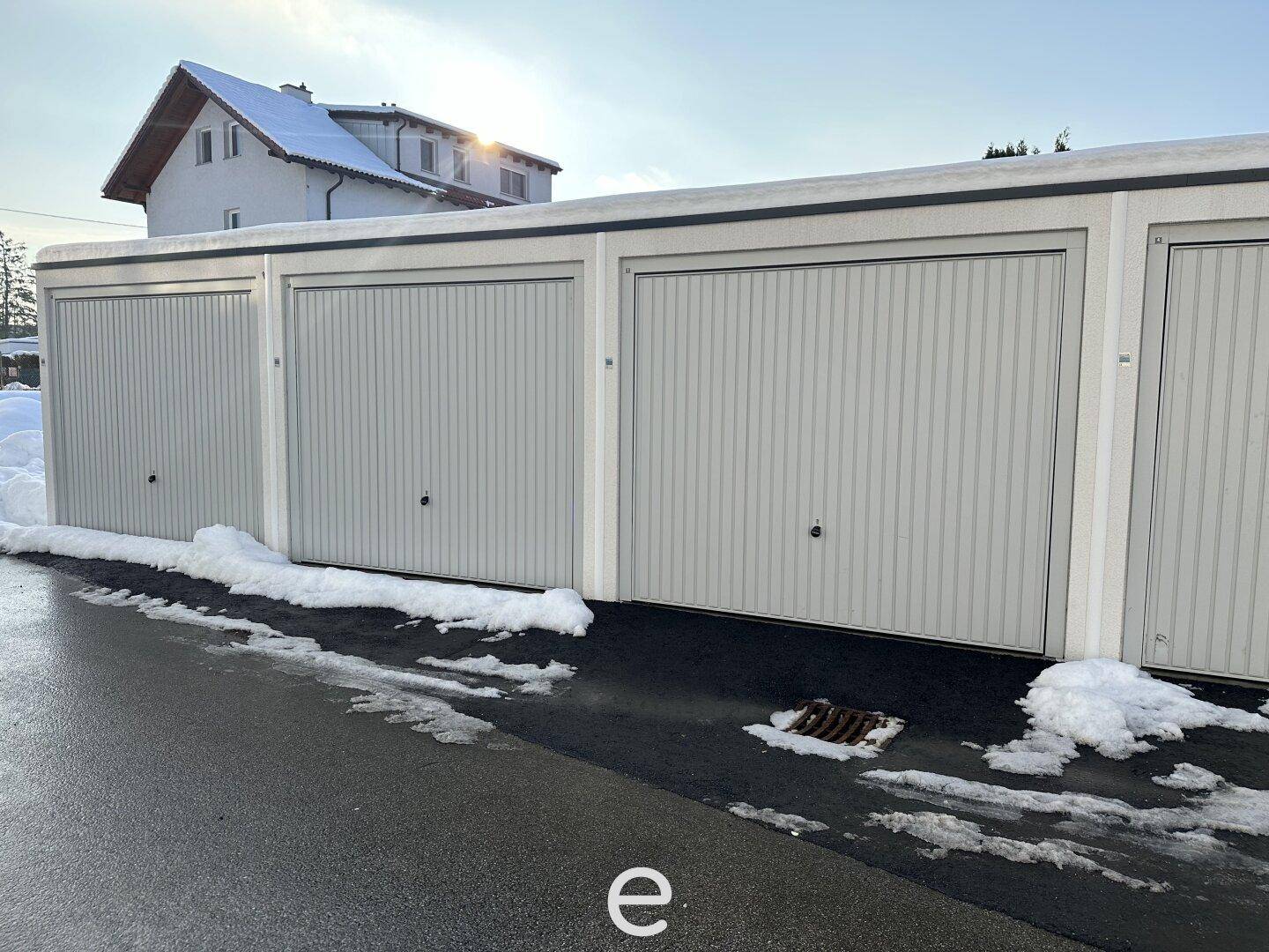 Reihe von Garagen mit Rolltoren, umgeben von Schnee und einem Wohnhaus im Hintergrund.